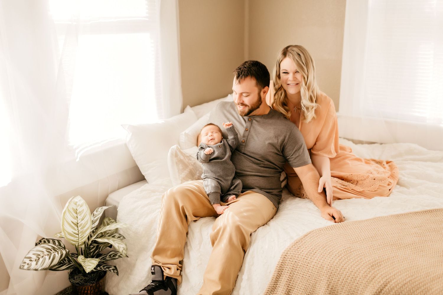 a mom and dad sit on a bed holding their baby and smiling while posing for a photo
