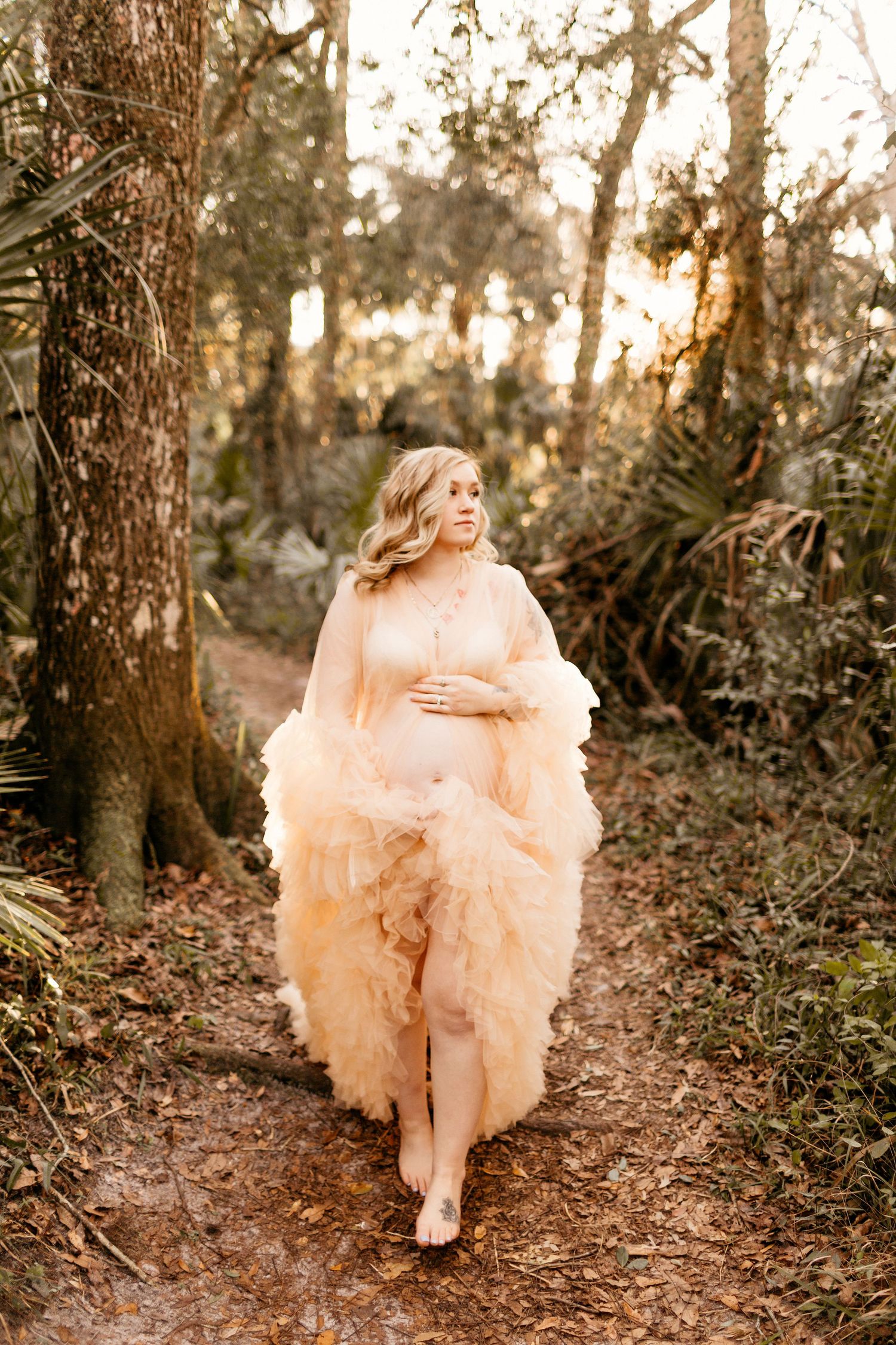 a pregnant mom walks through the woods holding her belly and looking to the side while posing for a photo