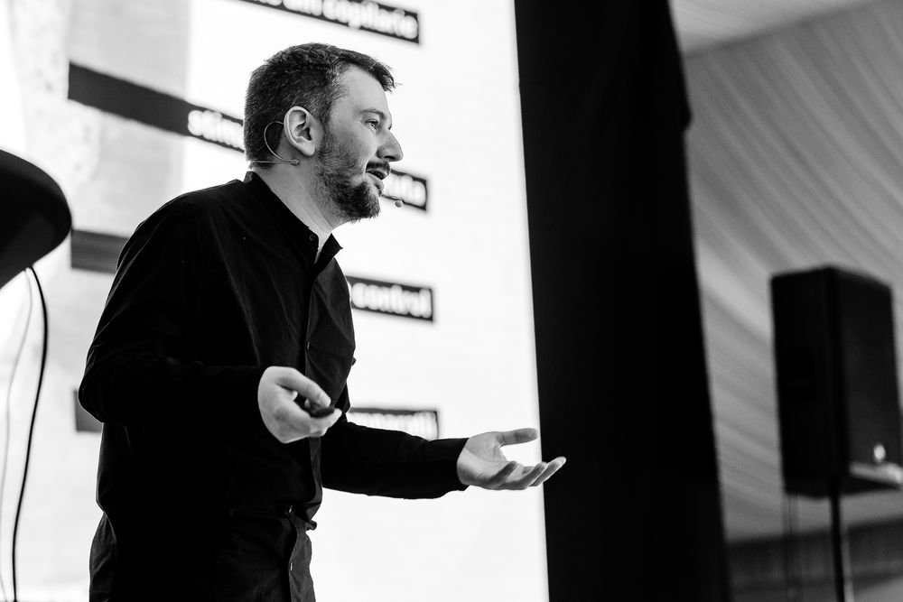 Speaker in black attire gesturing expressively during presentation in monochrome.