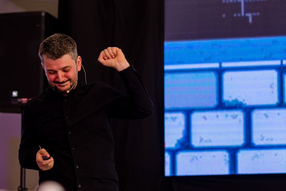 Energetic presenter in dark clothing against digital display background.