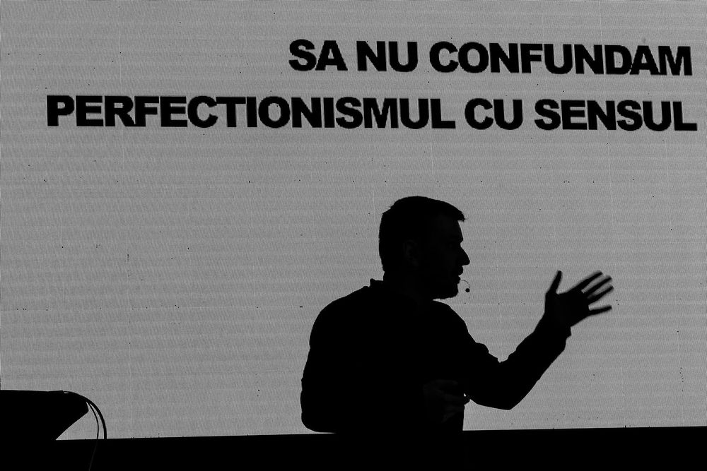 Silhouetted speaker gesturing against Romanian text projection backdrop.