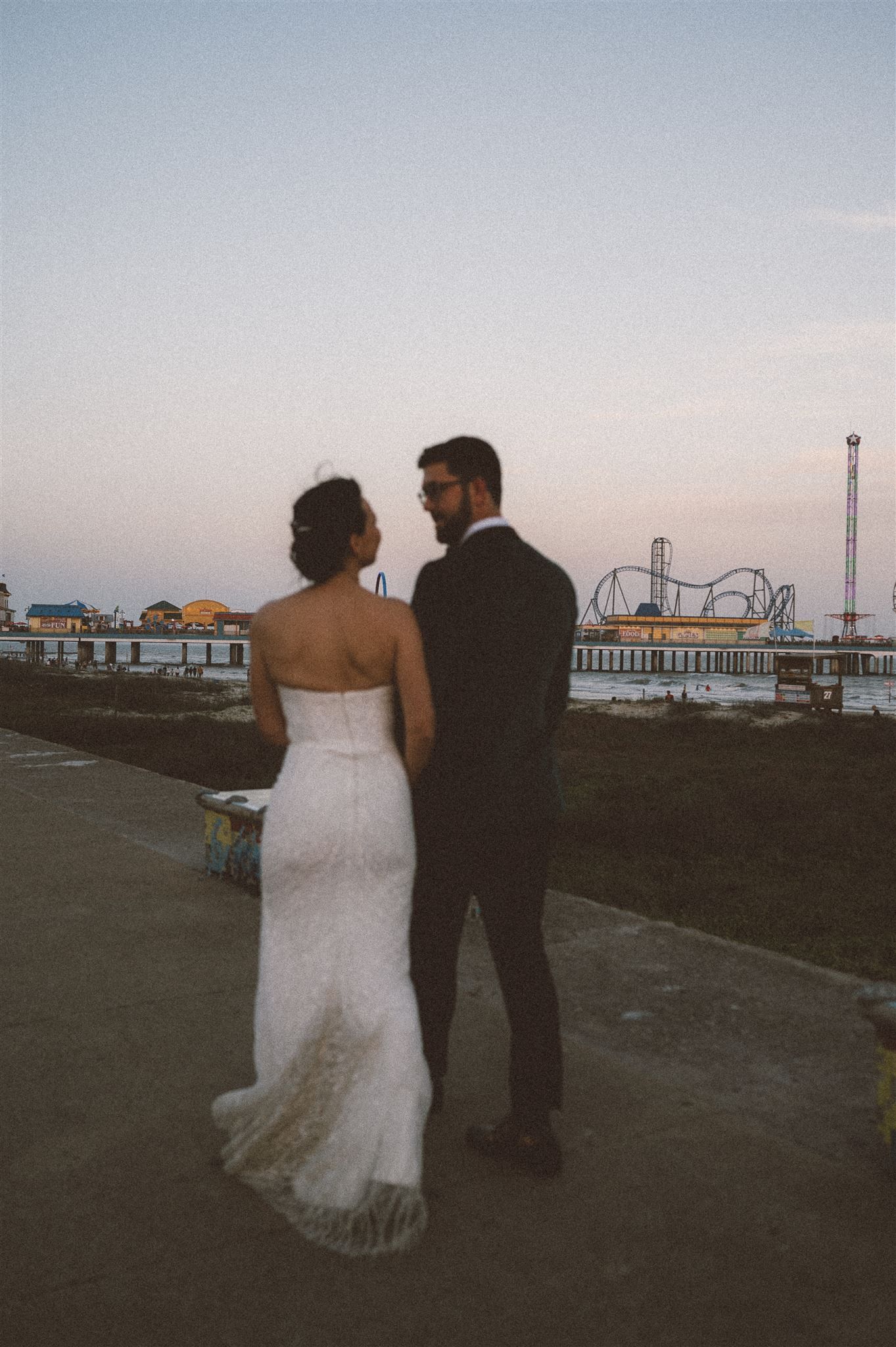 Silhouetted wedding couple stands together against sunset and pier backdrop.