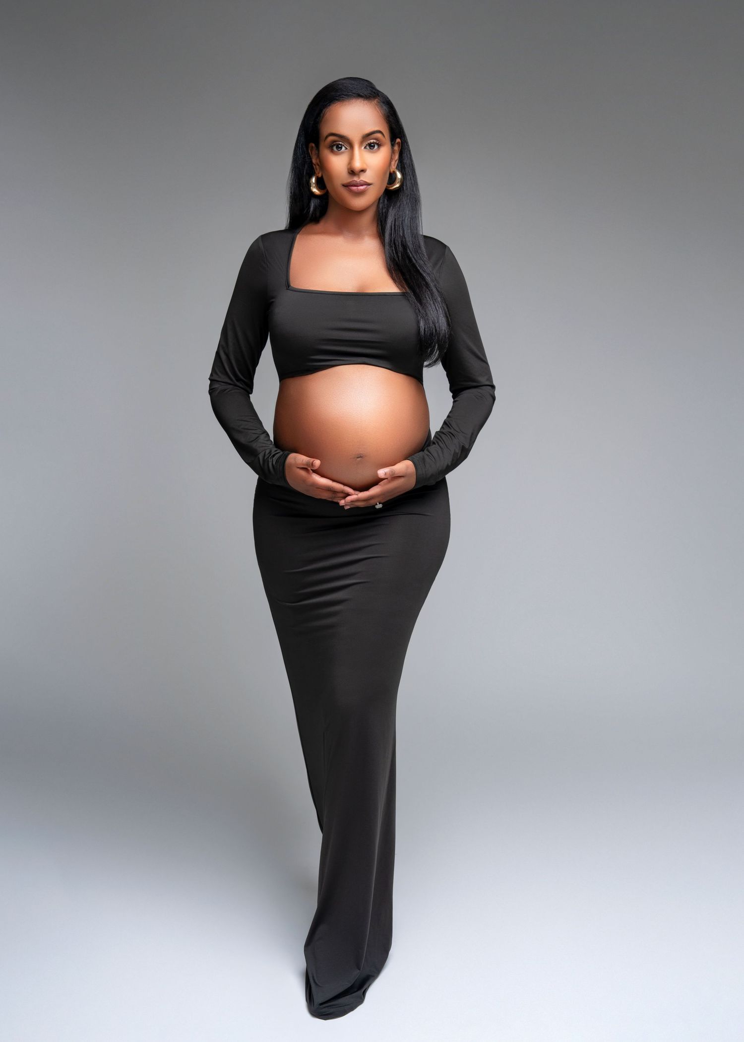 studio maternity photo shoot in an editorial style a woman stands with her hands under her belly and a soft smile