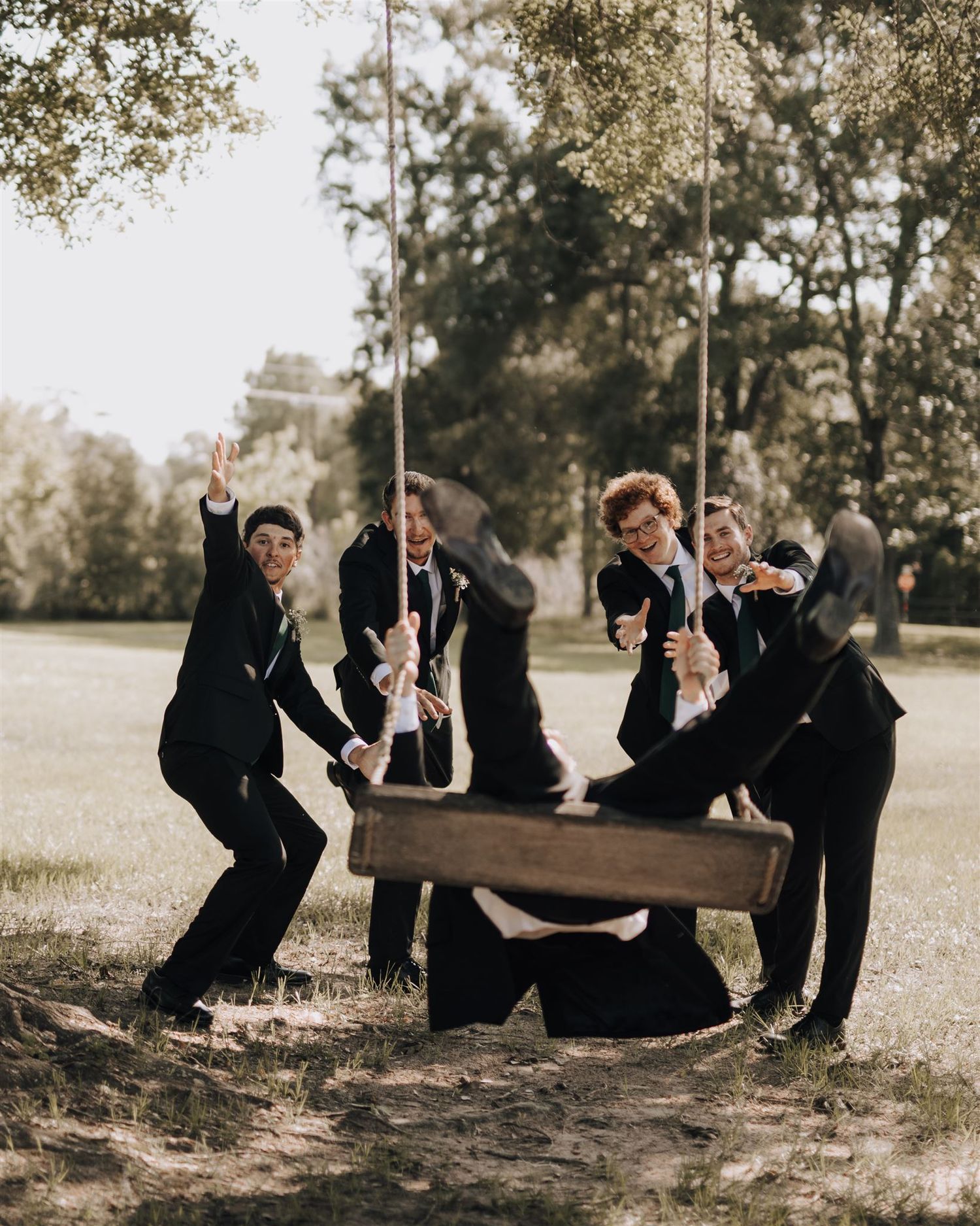 Wedding party members playfully pose around a wooden swing in a natural outdoor setting.