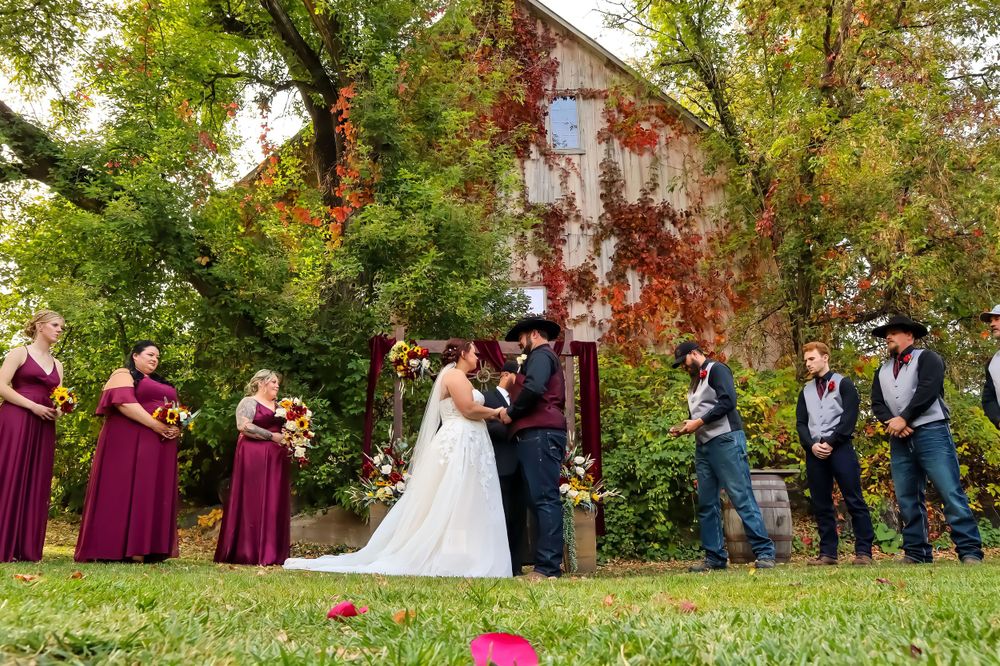 A wedding party poses outdoors with bridesmaids in burgundy and groomsmen in blue vests by a rustic barn.