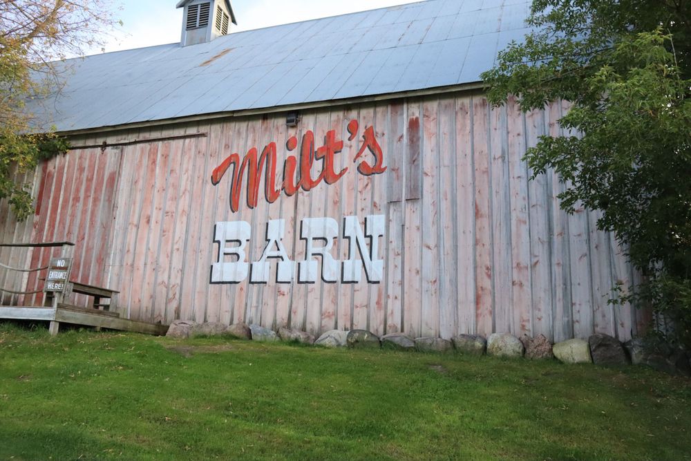 A weathered wooden barn with 'Milt's' painted on the side stands in a grassy field.