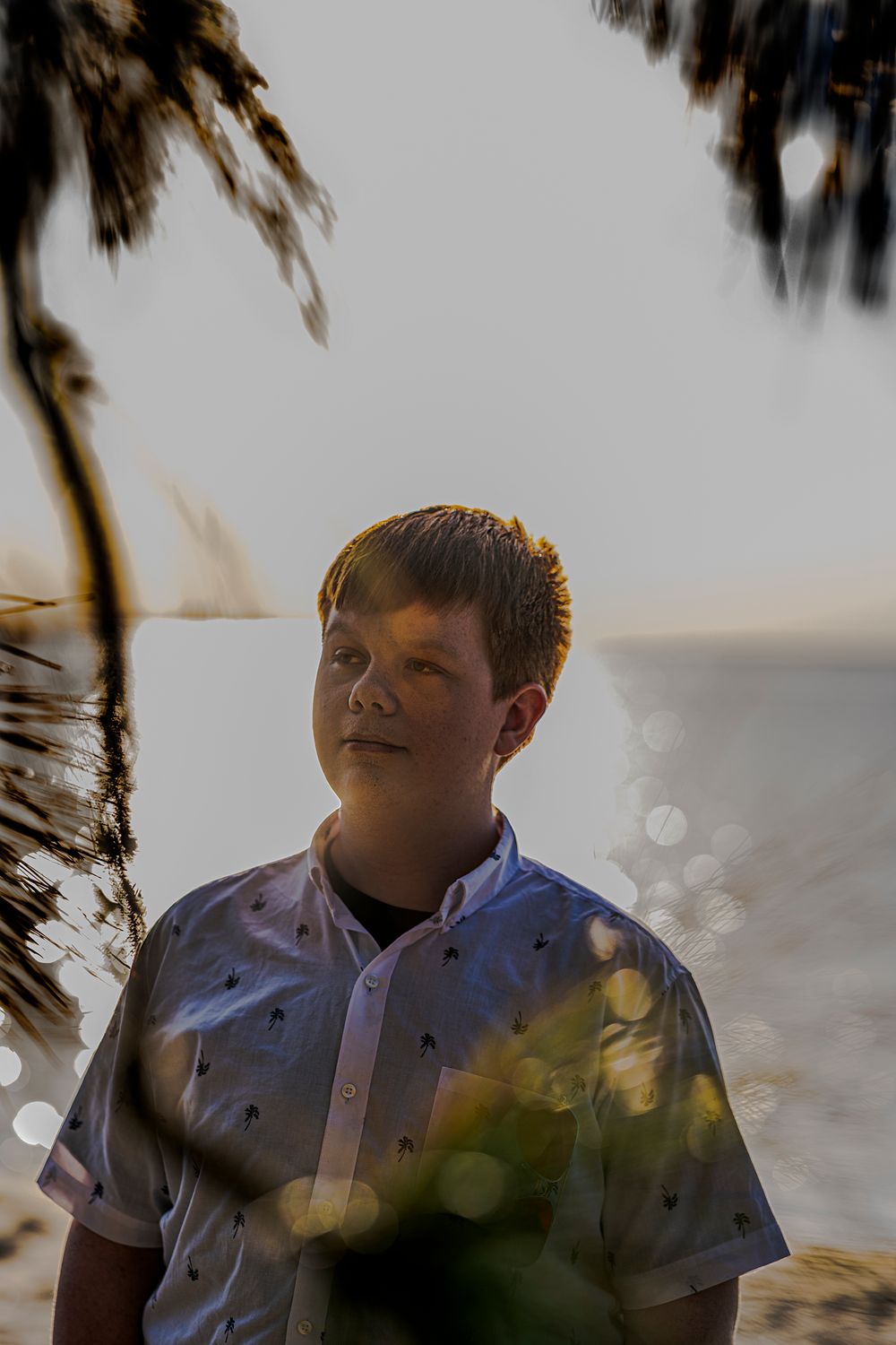 Portrait shot of youth in patterned shirt with palm trees and ocean in background.