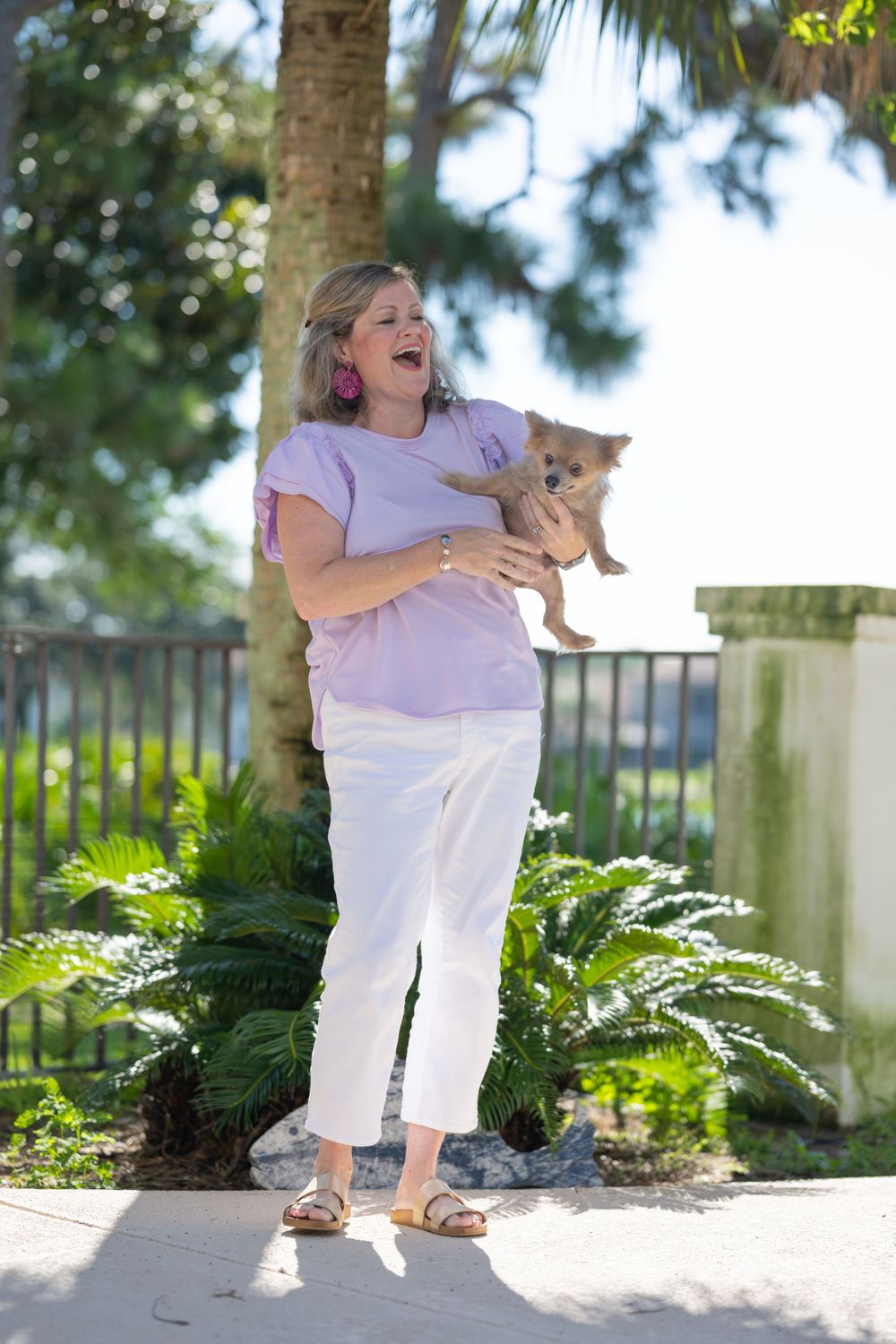 Person in pink shirt and white pants holding small dog while standing near palm trees.