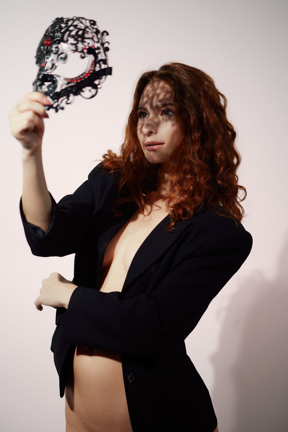 A person with curly red hair in a black blazer holds an ornate black masquerade mask against a light background.