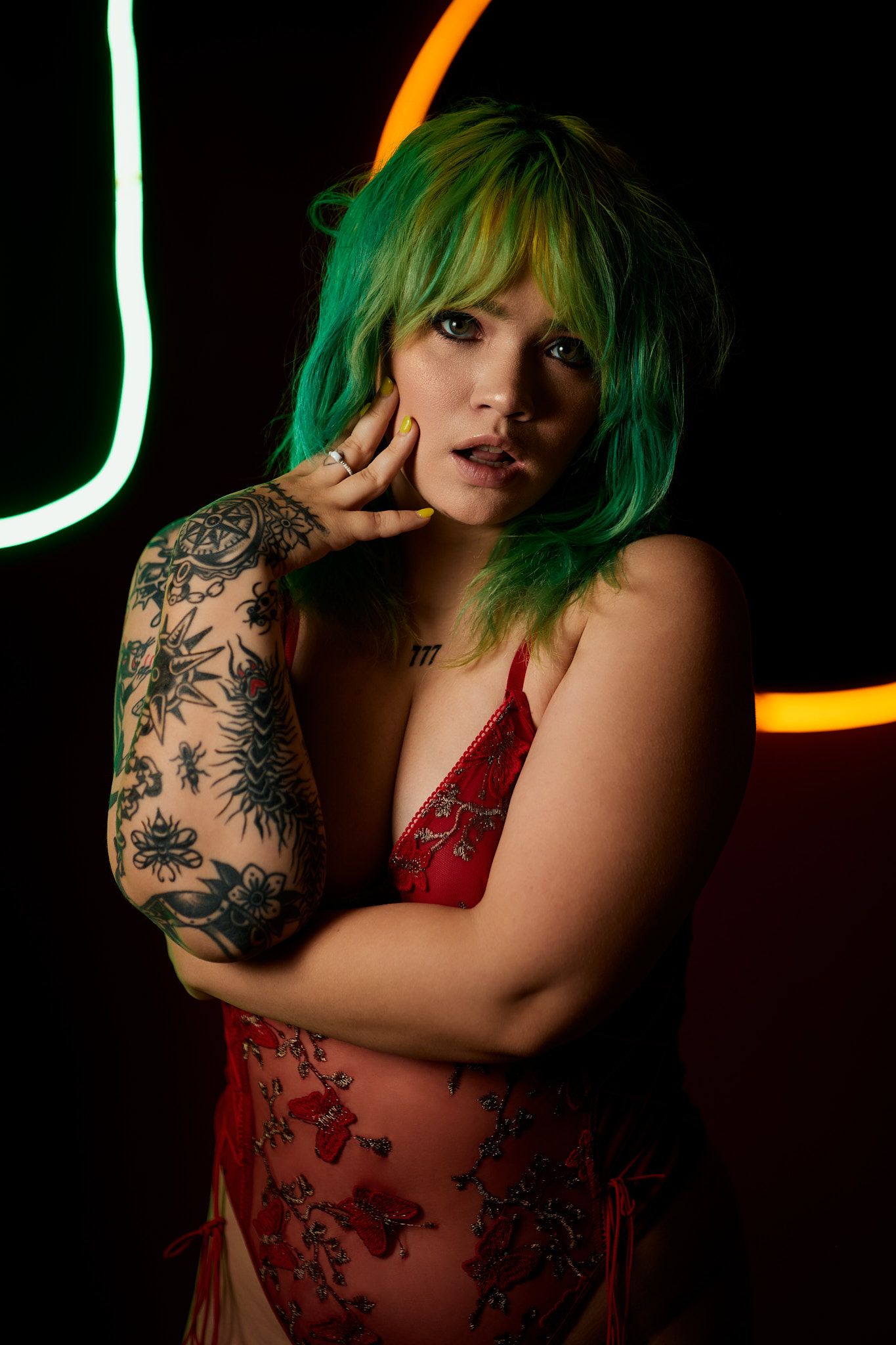 Artistic portrait of someone in red lace lingerie with green hair and floral arm tattoos in moody neon lighting.