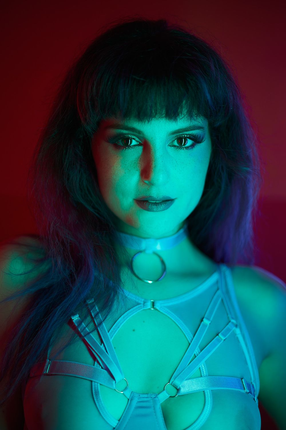 Artistic portrait in cyan and red lighting featuring dark hair and a metallic harness accessory.