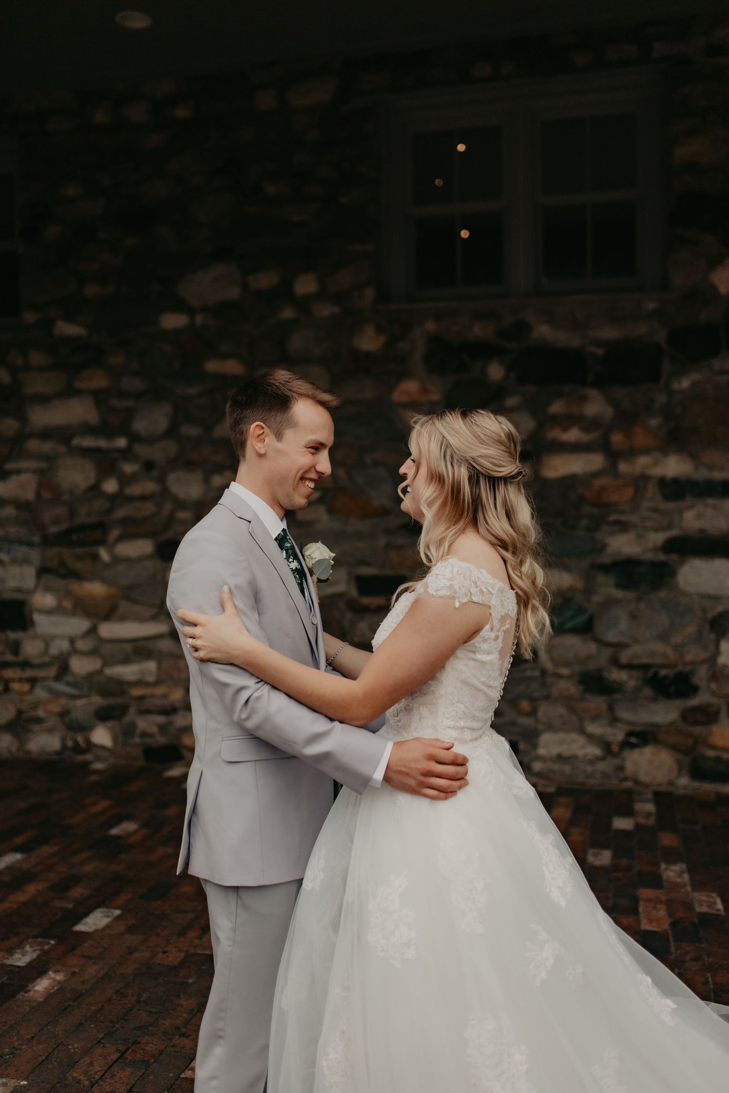 A couple shares a joyful moment together at their wedding, with the bride in white and groom in light gray against stone wall.
