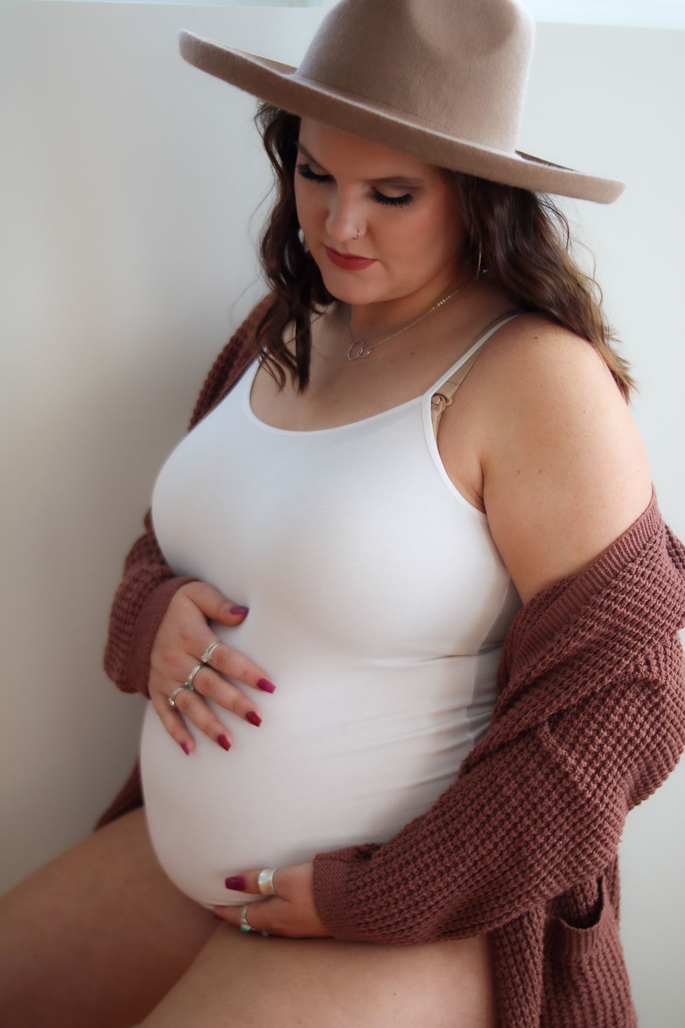 Person in white tank top and brown cardigan poses while holding their pregnant belly.