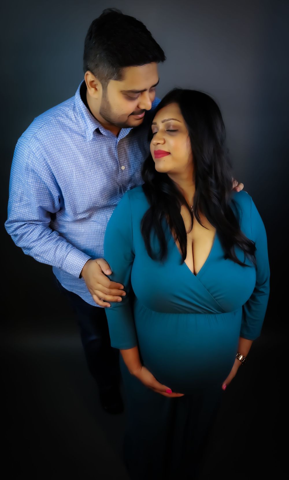 An intimate maternity photo of an expecting couple in blue and teal against a dark background.