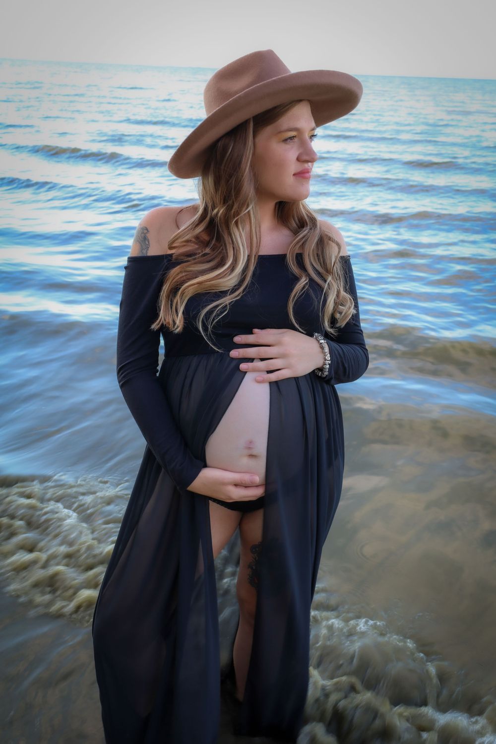 A maternity photo at the beach with waves and a brown hat.