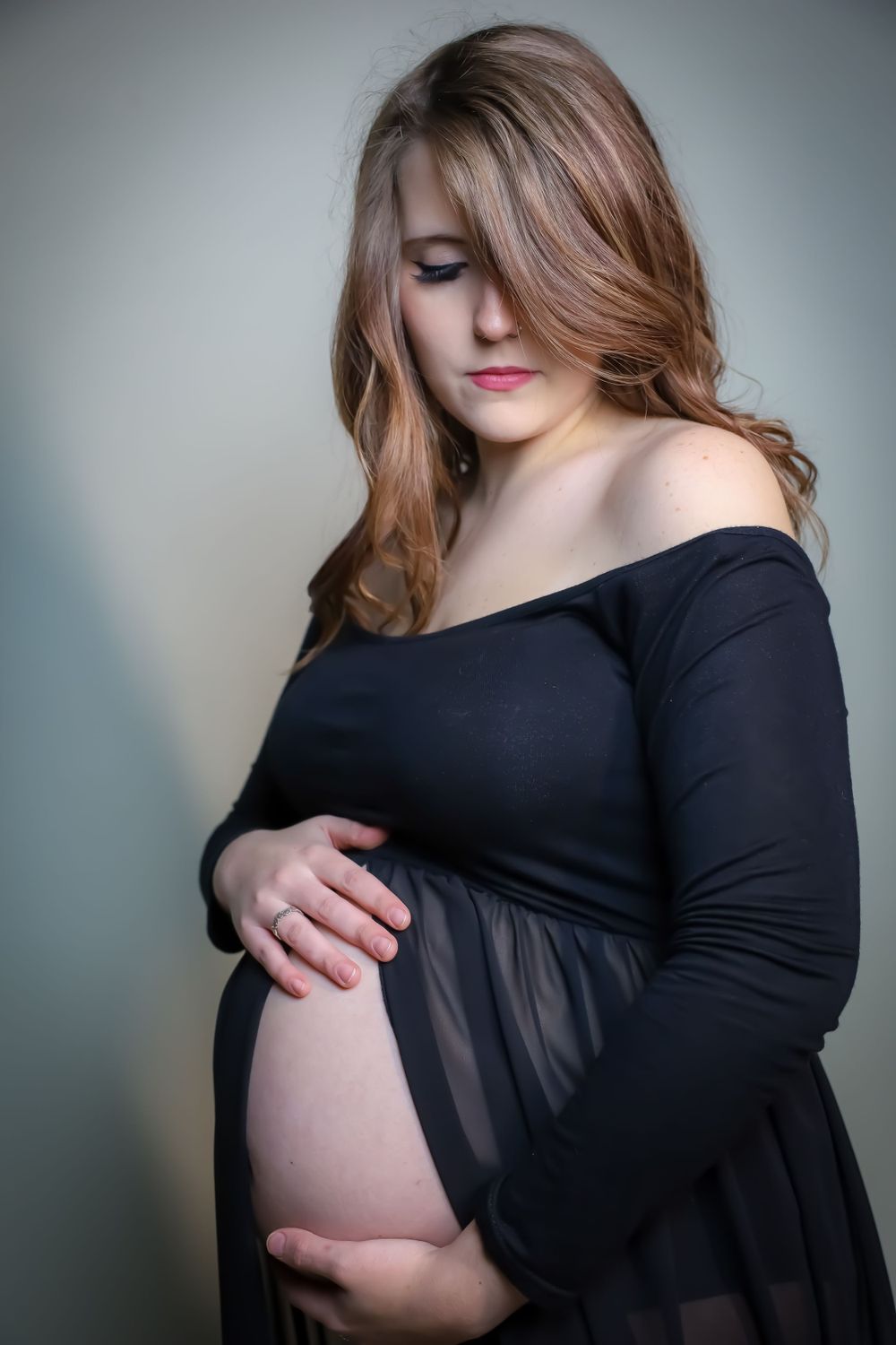 Woman in black maternity dress poses against grey backdrop for intimate photoshoot.