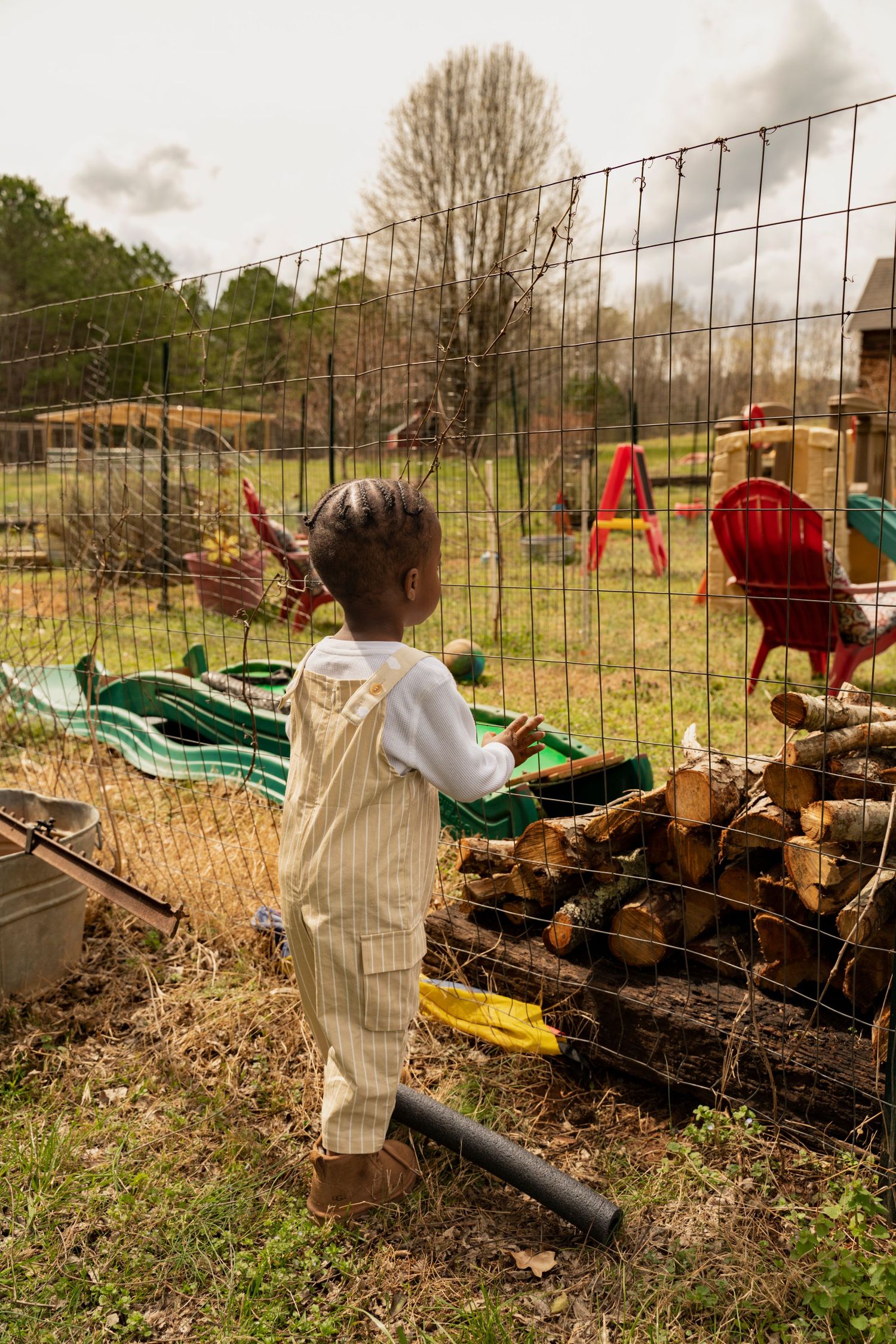 A small child in overalls looks through a wire fence at outdoor playground equipment.