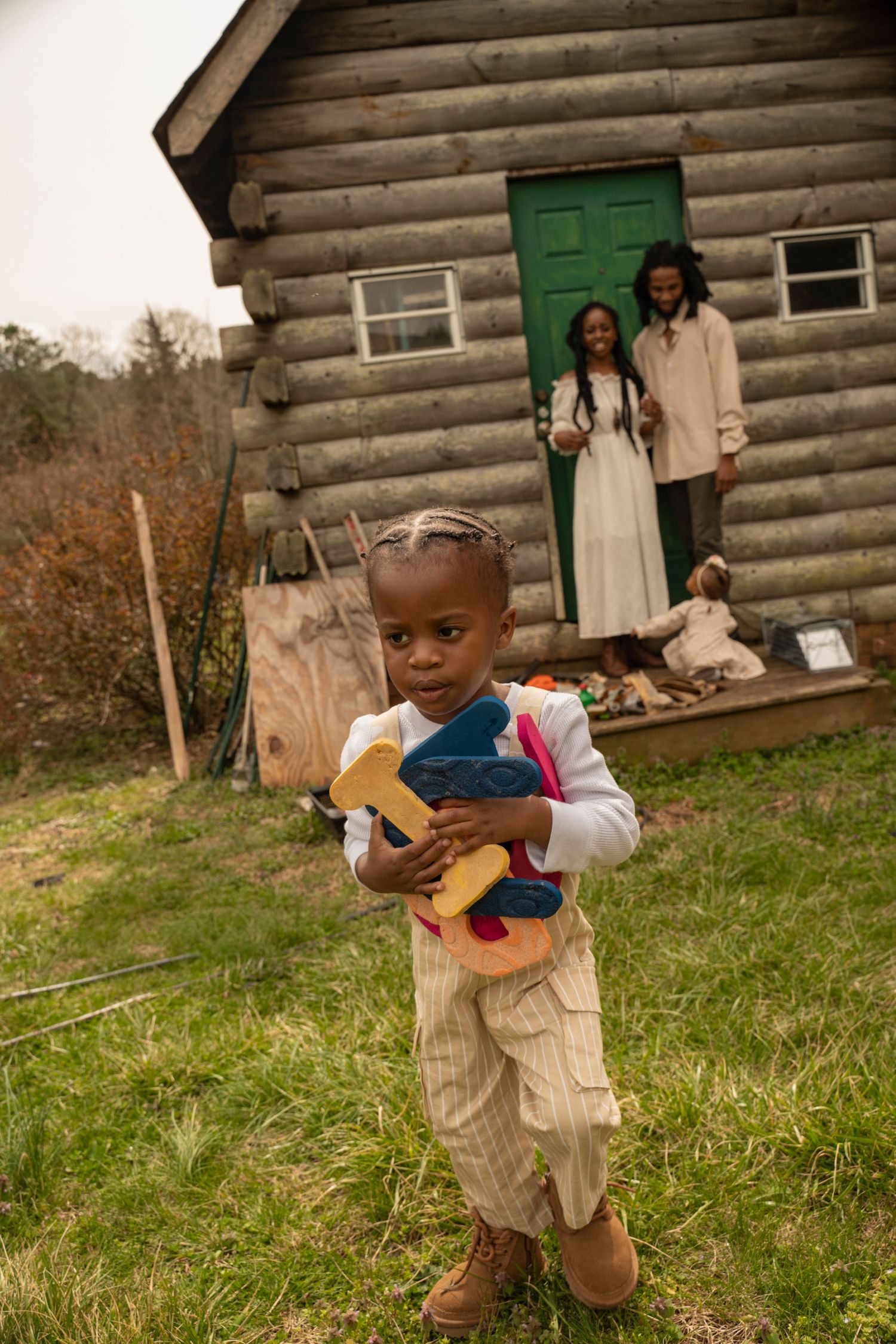 A child holds a toy while standing in front of a rustic log cabin with others in background.