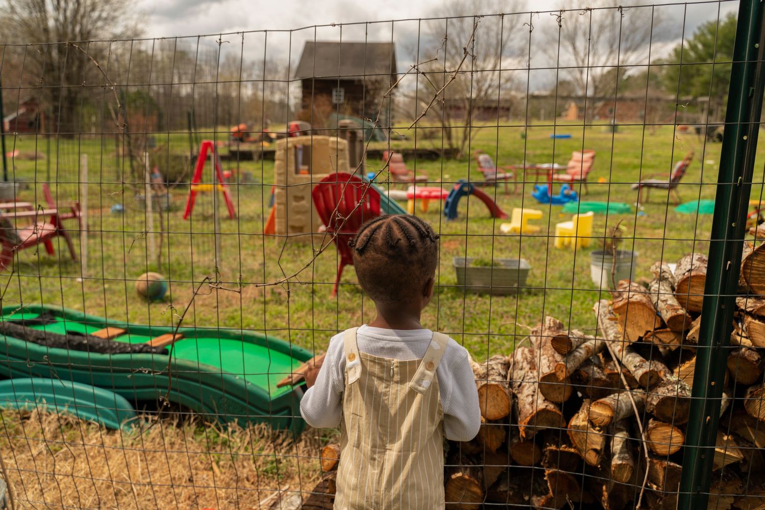 A view through a wire fence of a playground with colorful equipment and wood pile.