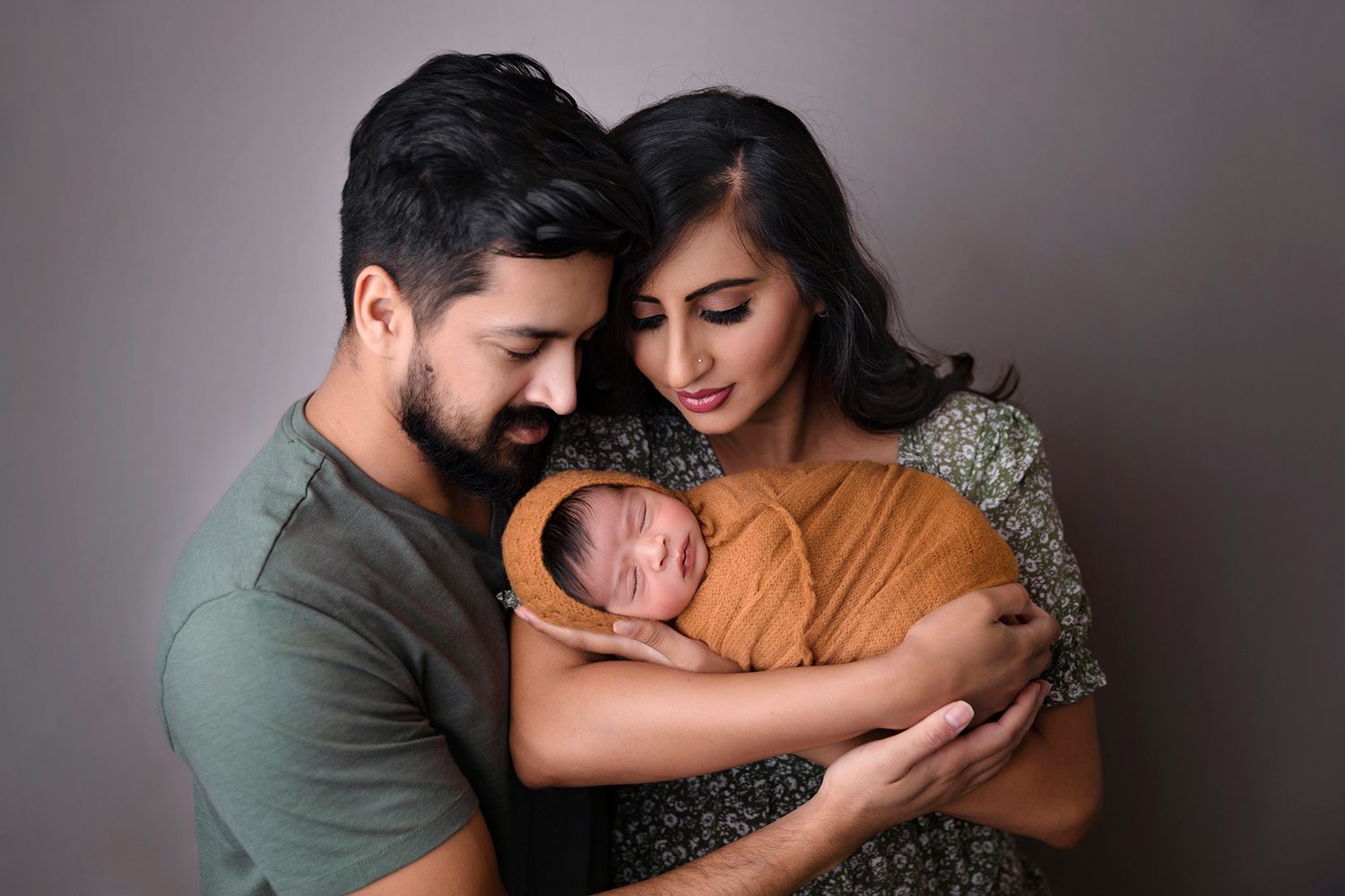A newborn baby swaddled in mustard yellow fabric being held lovingly against a gray backdrop.