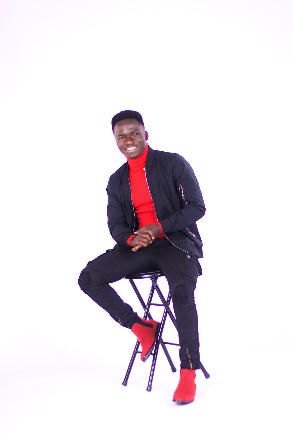 Smiling young man in black jacket and red turtleneck sits on stool indoors.