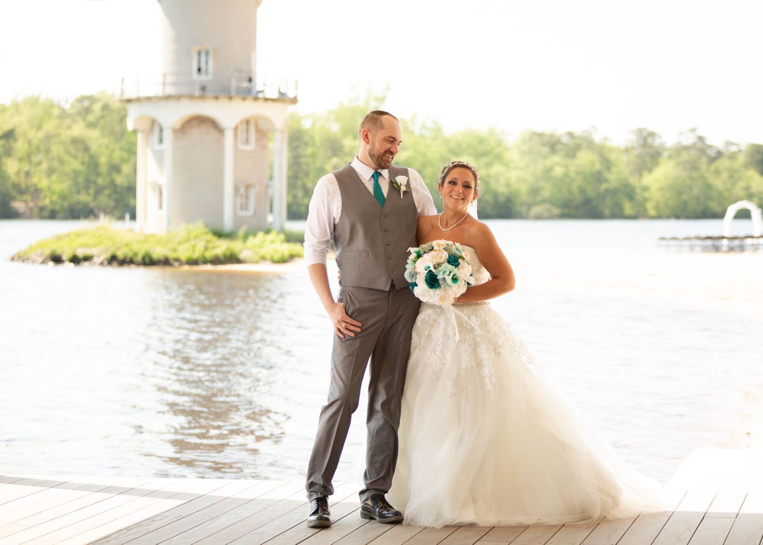 Bride and groom pose on a waterfront dock in elegant wedding attire with a tower in the background.