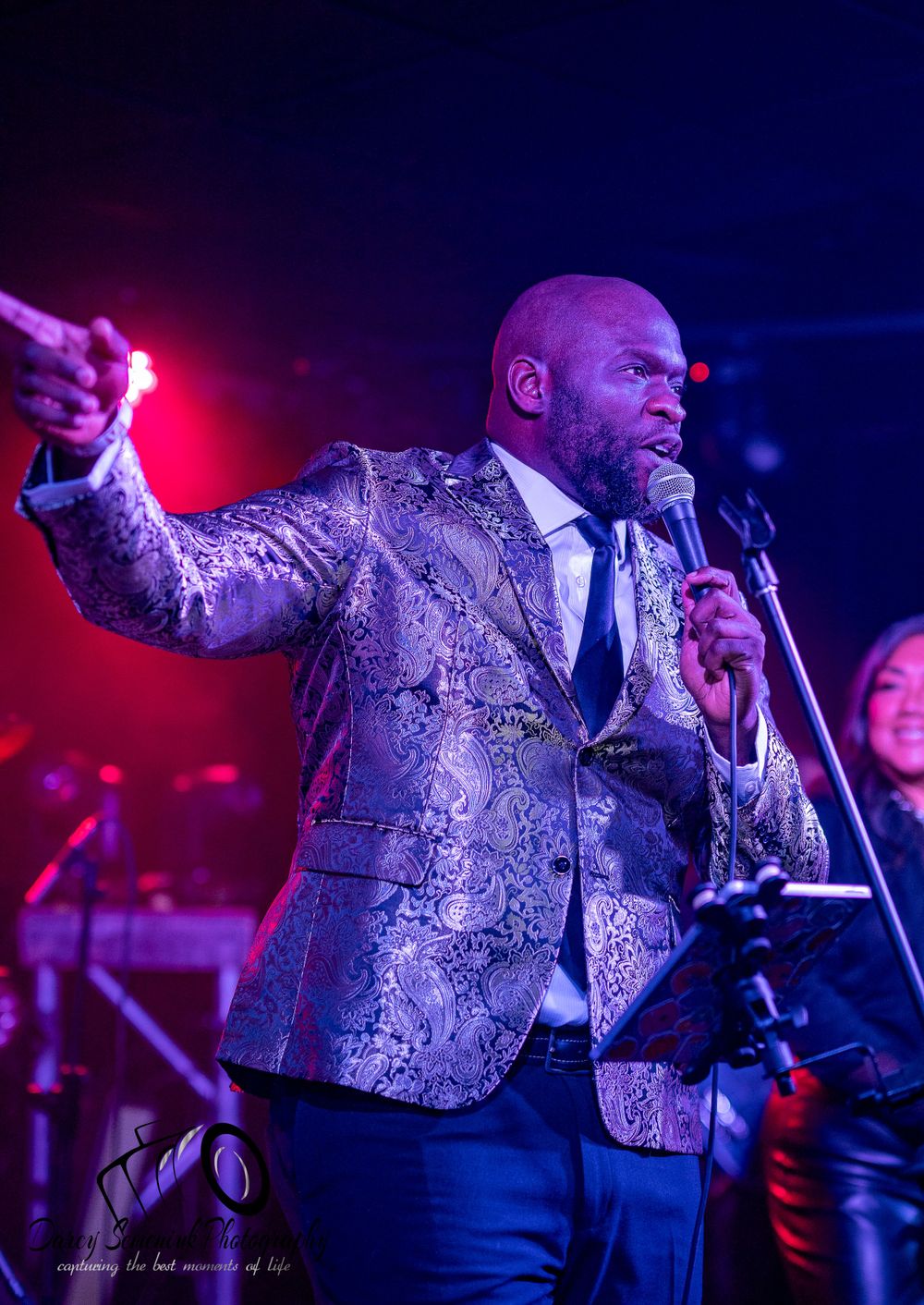 A performer in a shimmering silver patterned vest sings passionately on stage under dramatic purple and red lighting.