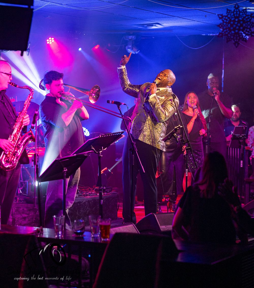 Musicians perform on stage with vibrant blue and pink lighting creating an energetic concert atmosphere.