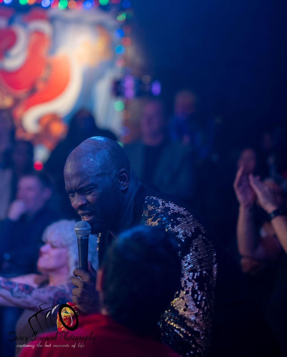 A person sits in a dimly lit nightclub with colorful bokeh lights creating an atmospheric ambiance in the background.