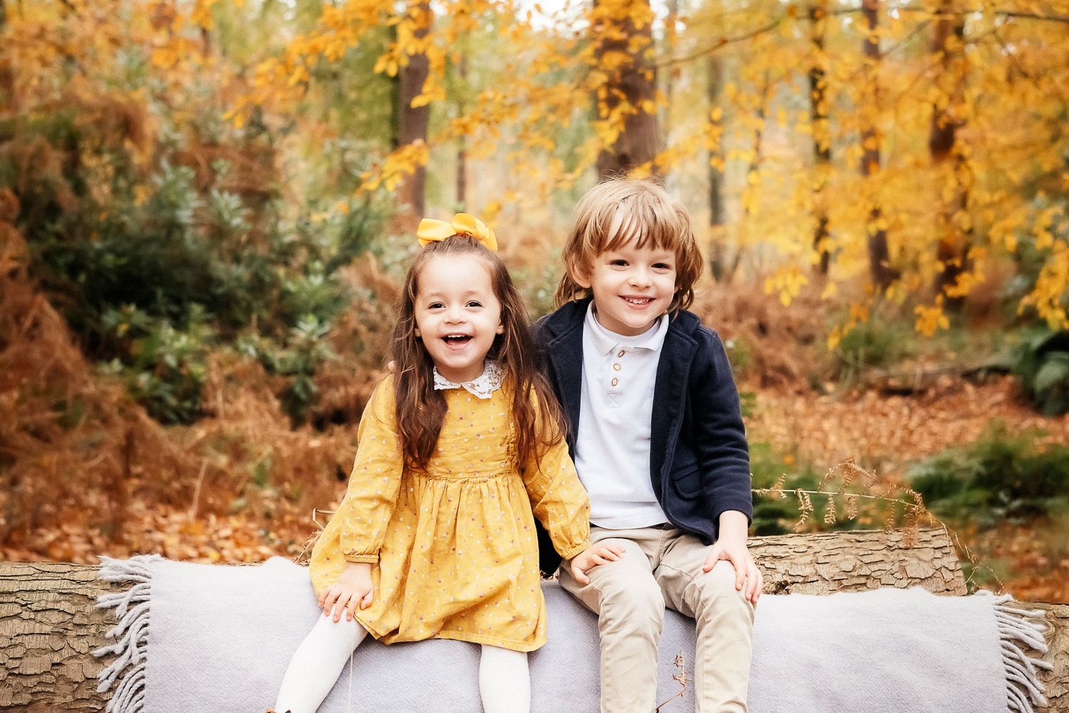 Two children sitting together on a bench surrounded by golden autumn foliage.