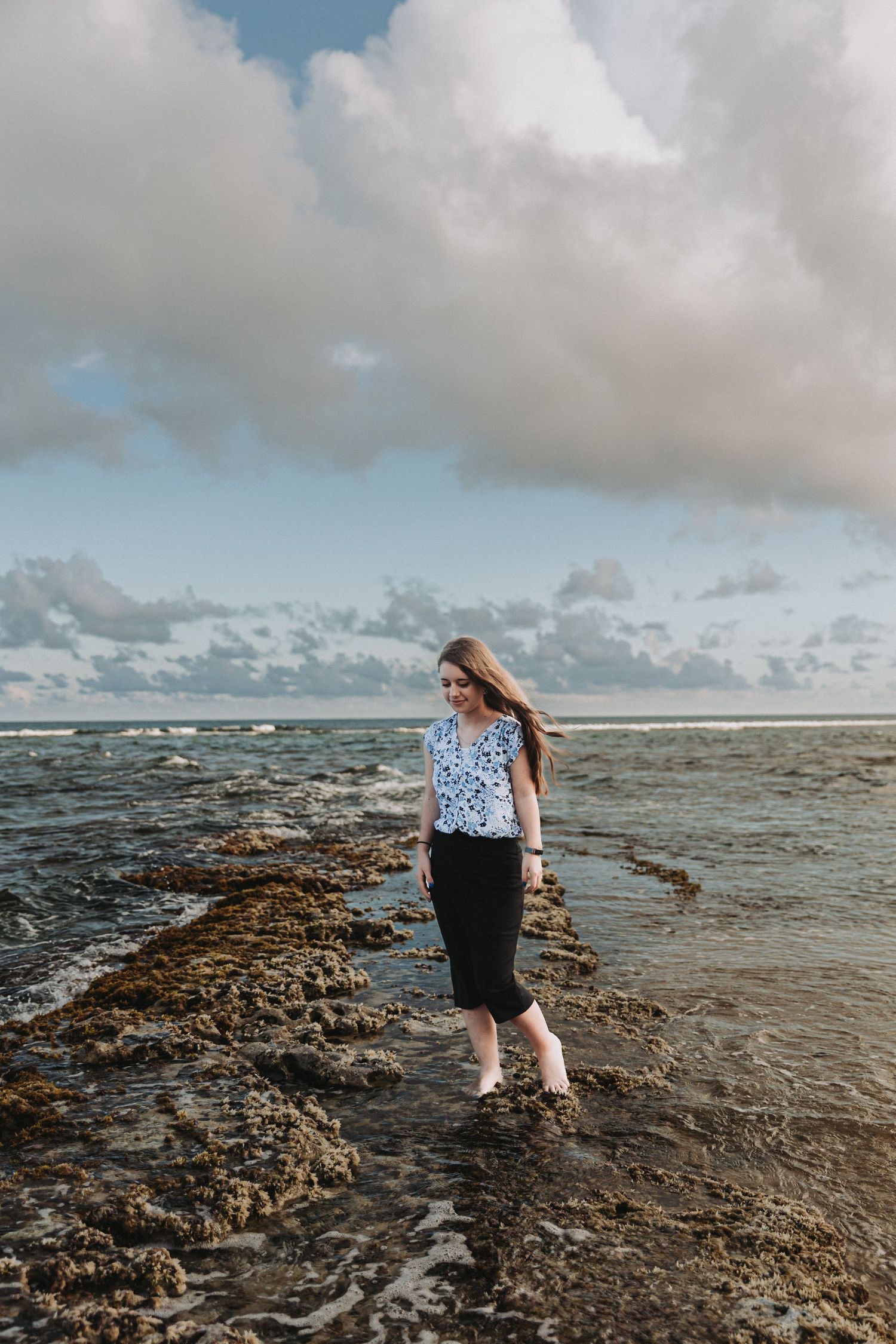 A person stands on rocky ocean shore wearing a floral top and black skirt as clouds gather overhead.