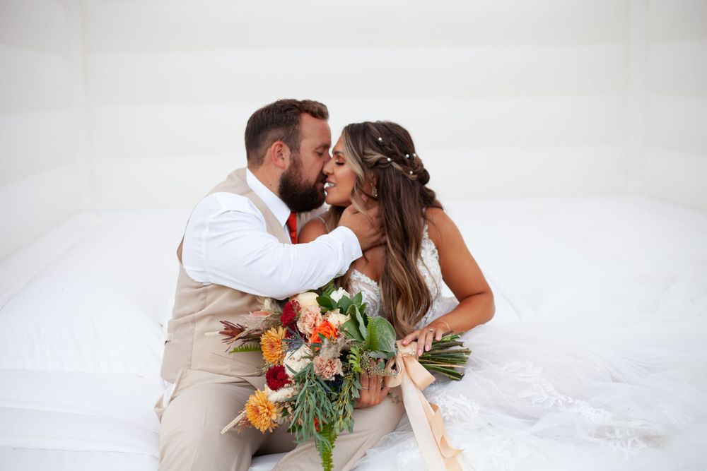Romantic wedding couple shares a tender kiss while holding a vibrant orange and green bouquet against white bounce house.