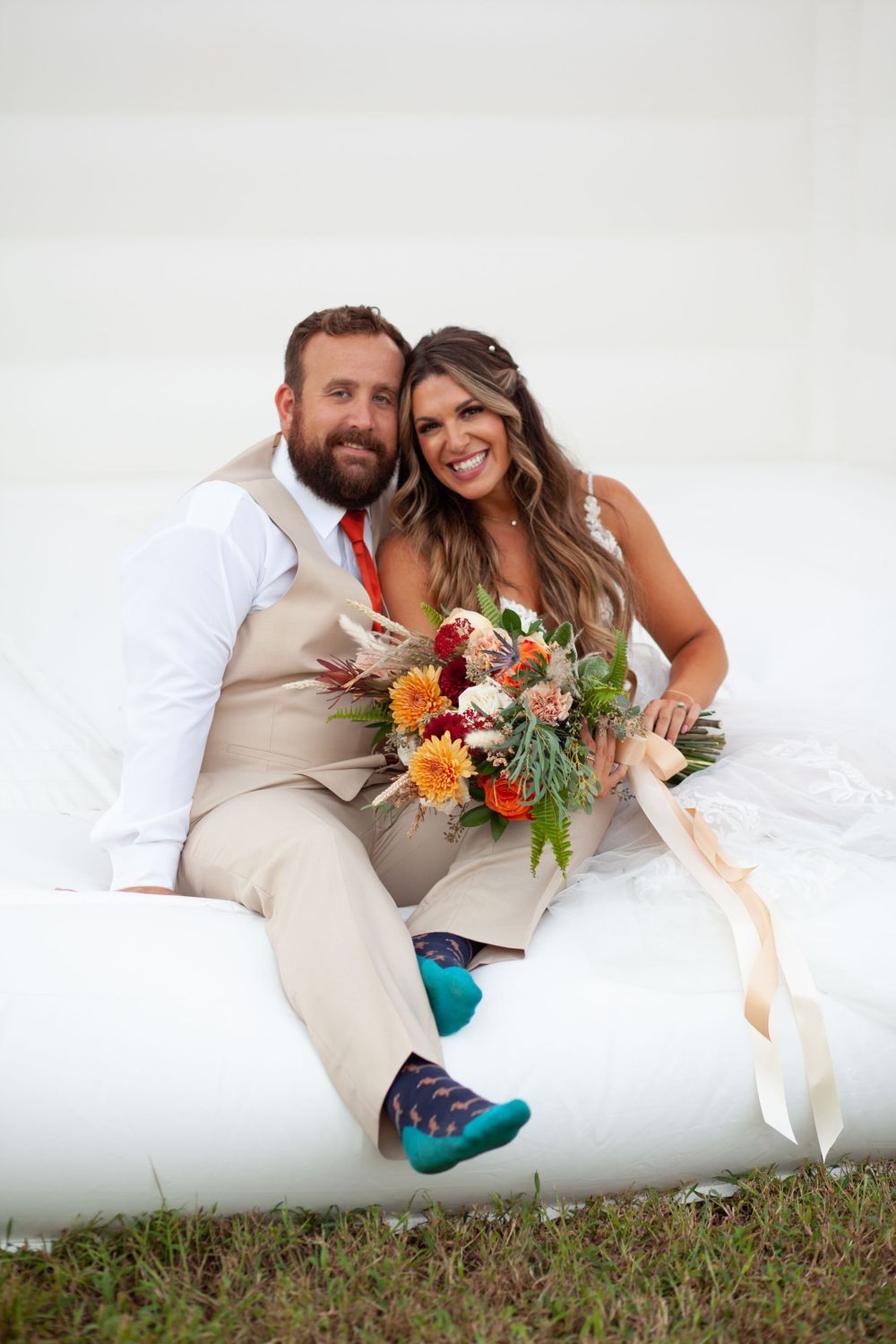 Newlyweds sit together on white bounchouse outdoors with vibrant orange and pink floral bouquet