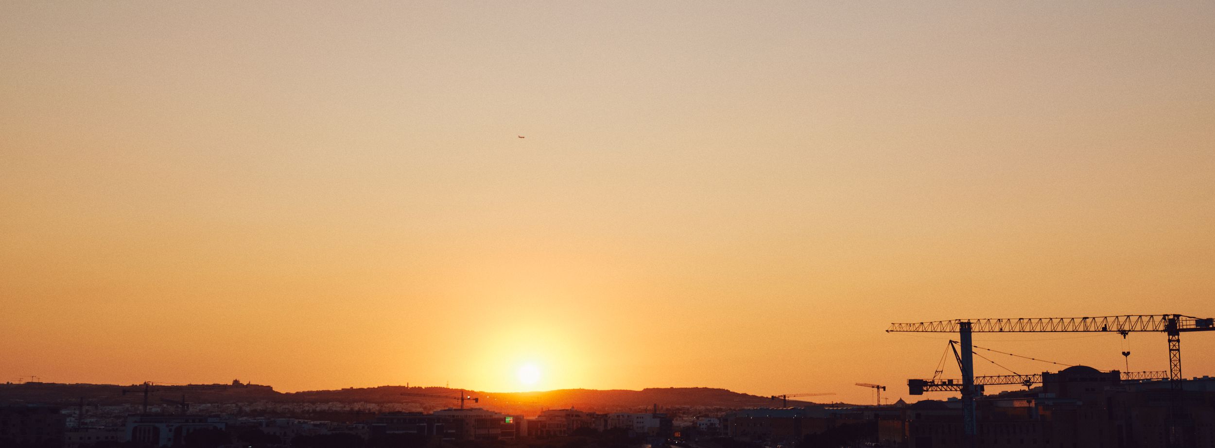 Silhouette of a construction crane against a sunset sky over a city skyline.