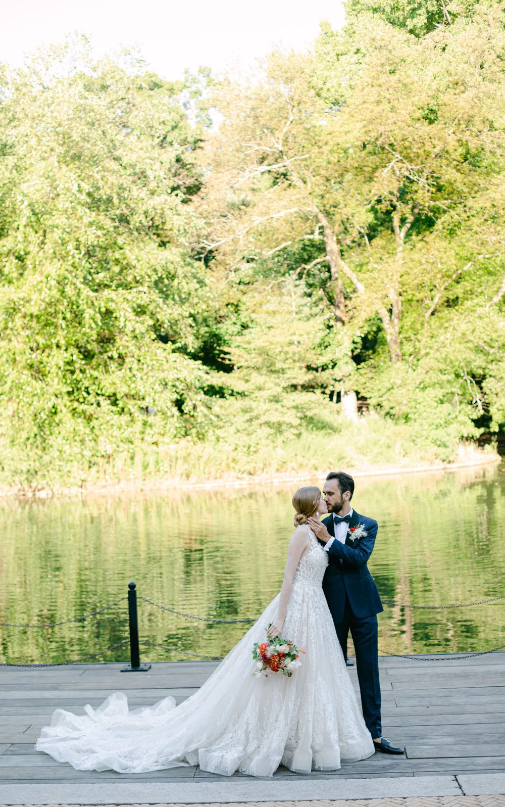Wedding couple shares intimate moment on wooden deck overlooking serene lake and lush trees.