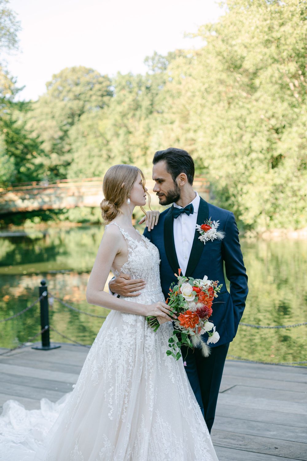 Bride in lace gown and groom in navy suit pose with coral bouquet by lakeside.