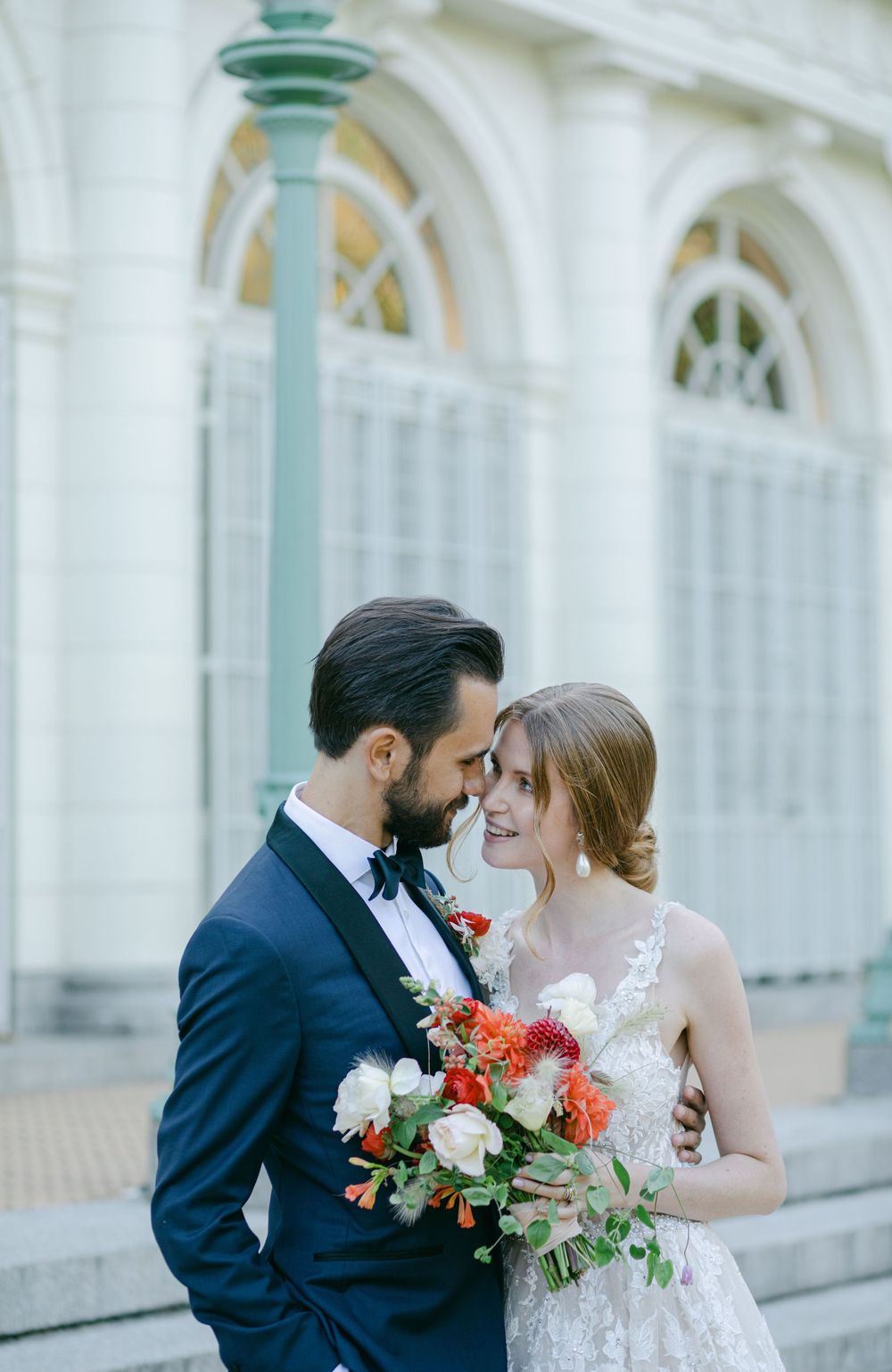 A romantic wedding couple shares an intimate moment against a white columned building with a vibrant coral bouquet.