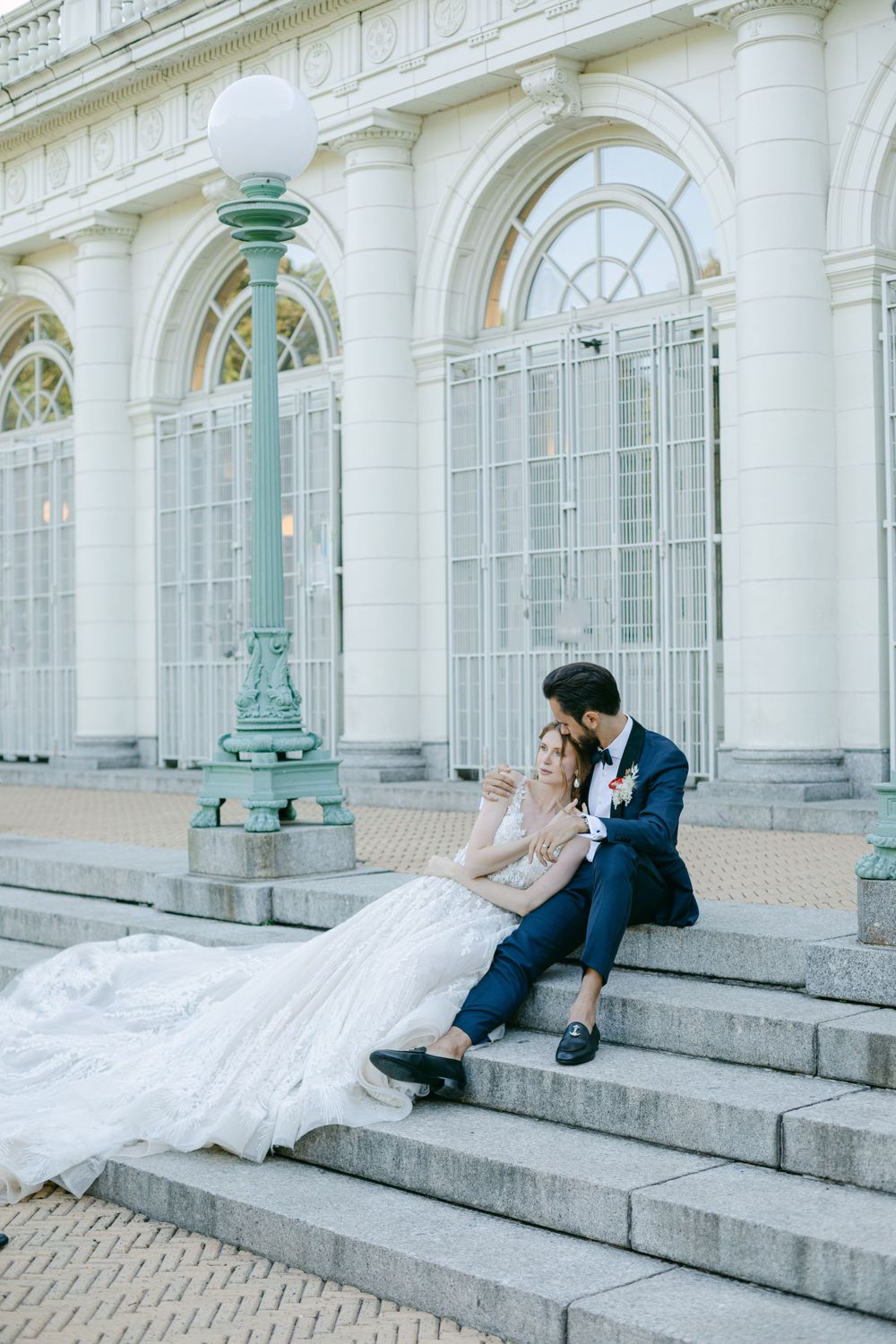 A couple sits on grand stone steps against classic white architecture with mint green lamposts.