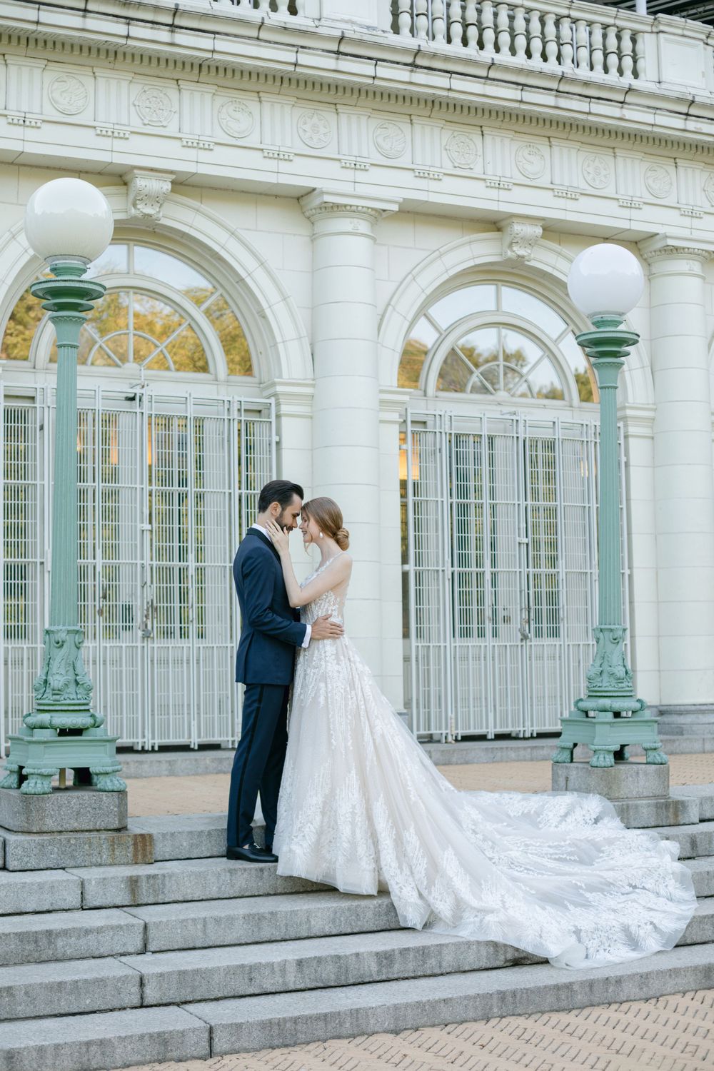 Wedding couple embraces on marble steps of classical building with ornate lamp posts and arched windows.