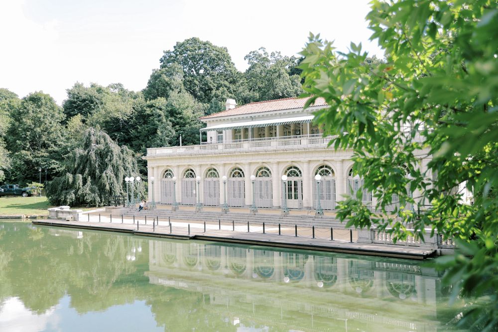 White neoclassical building with arched windows reflected in a tranquil pond surrounded by greenery.