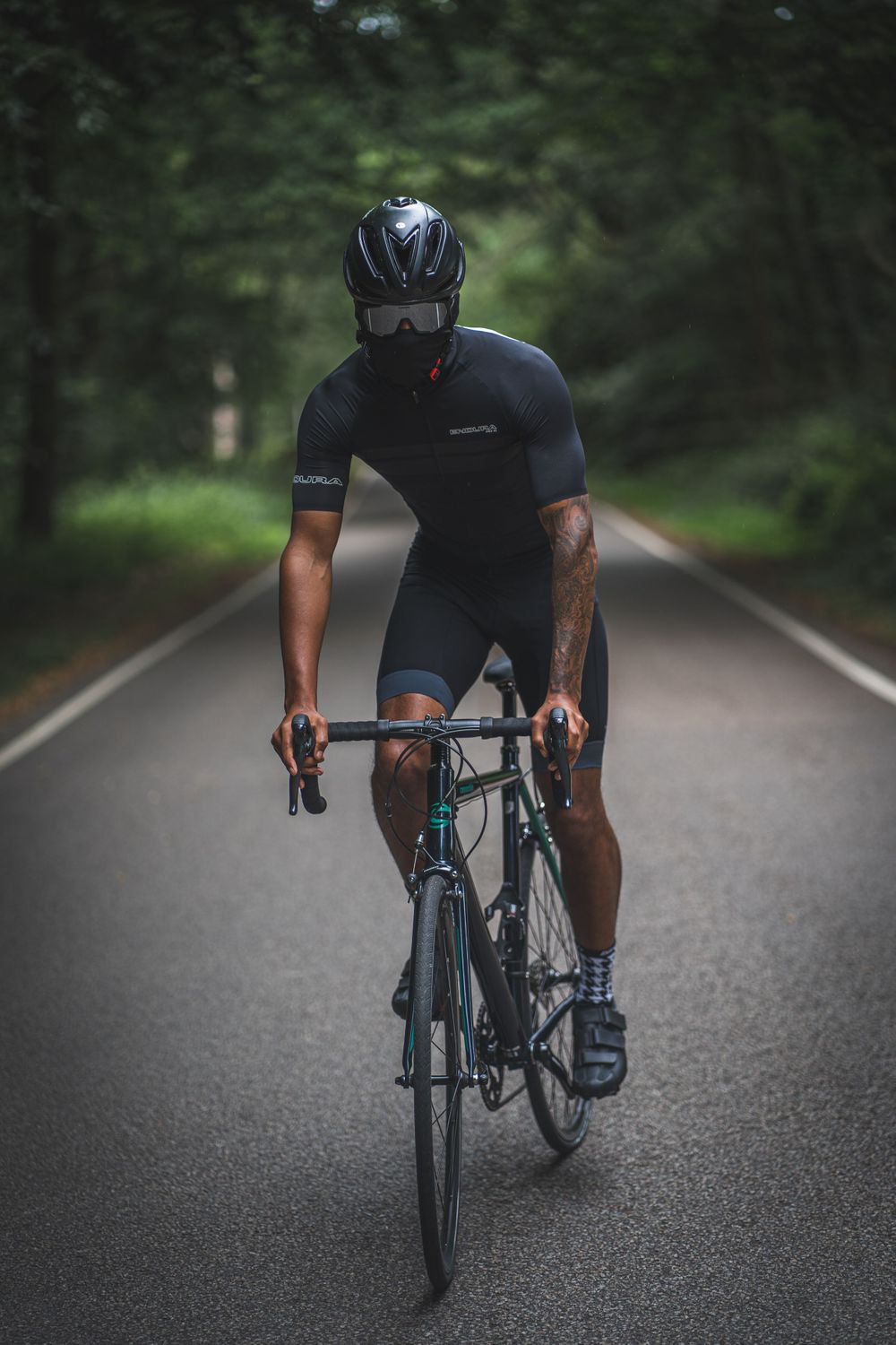 Cyclist in dark sportswear rides road bike on tree-lined path.