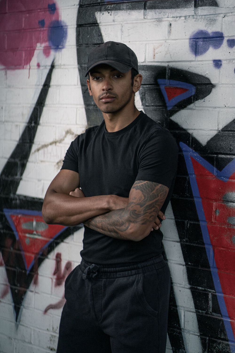 Athletic figure in black attire with tattoos stands against graffiti wall in urban setting.