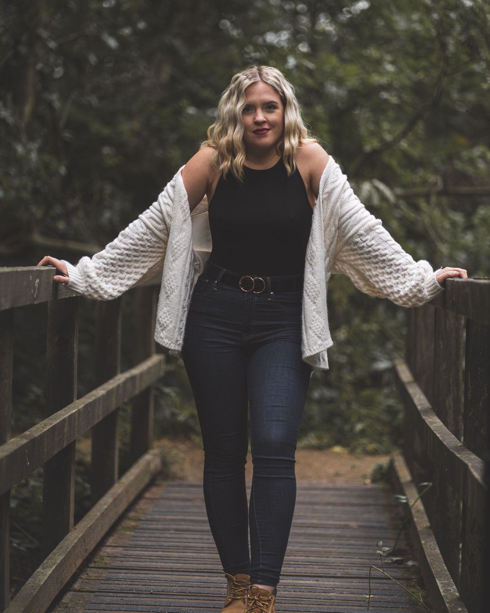 A person wearing white cardigan and black top poses on wooden bridge in forest setting.