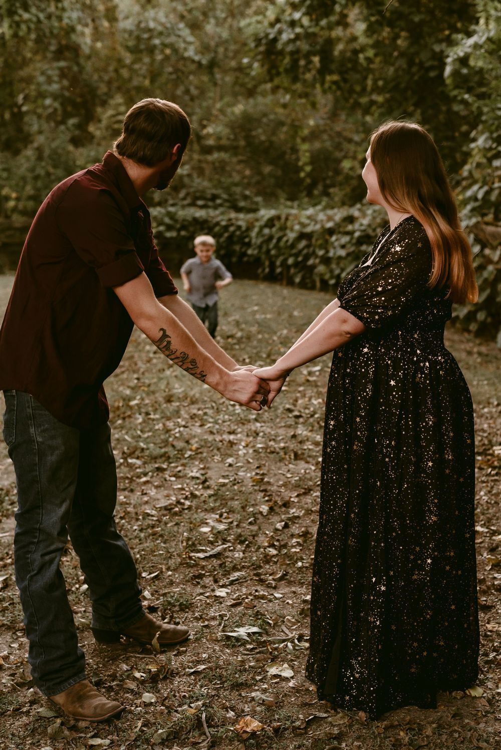 A couple holds hands while their child runs in the background of a wooded area.