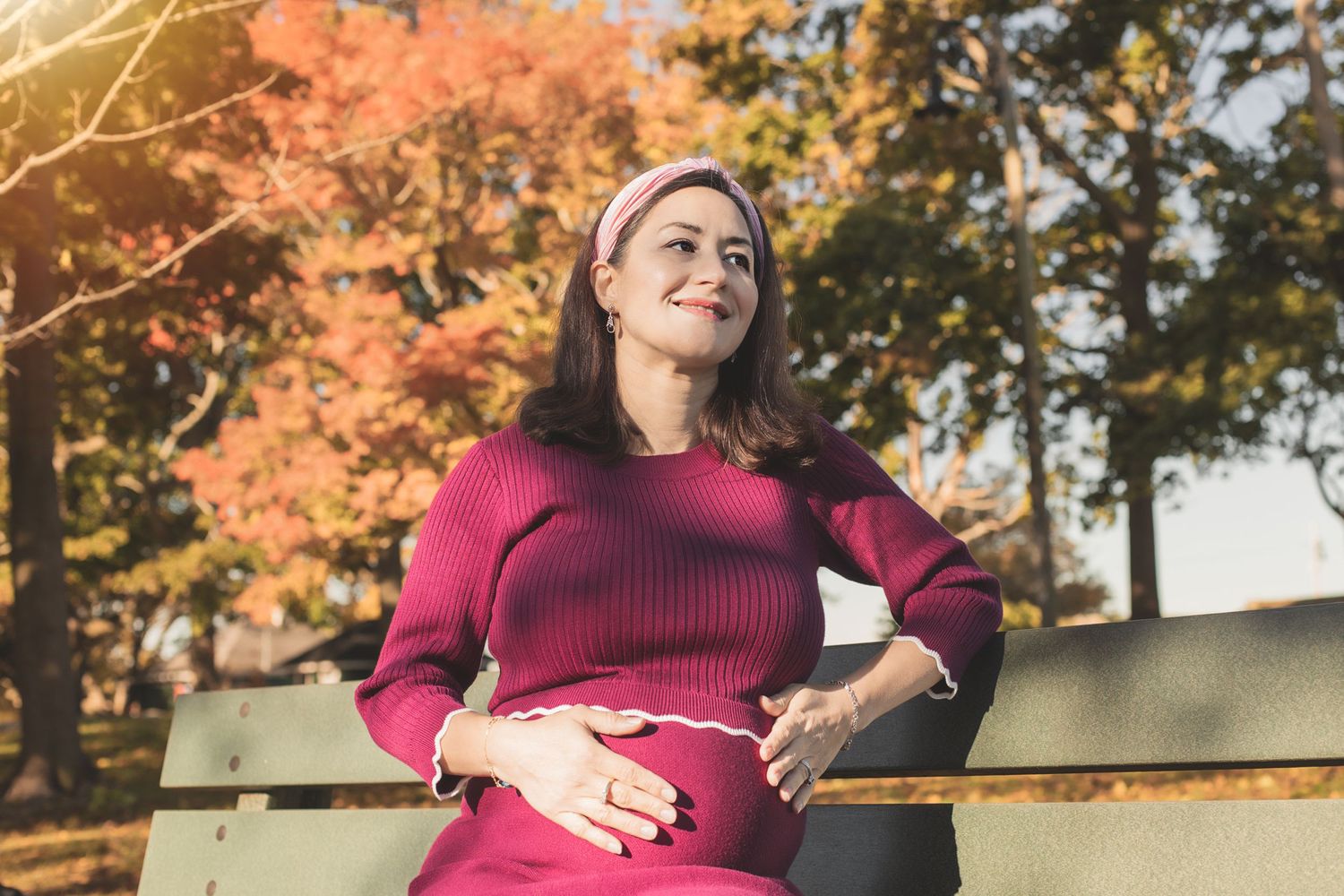 Pregnant woman in red sweater sitting on bench during autumn with orange leaves in background.