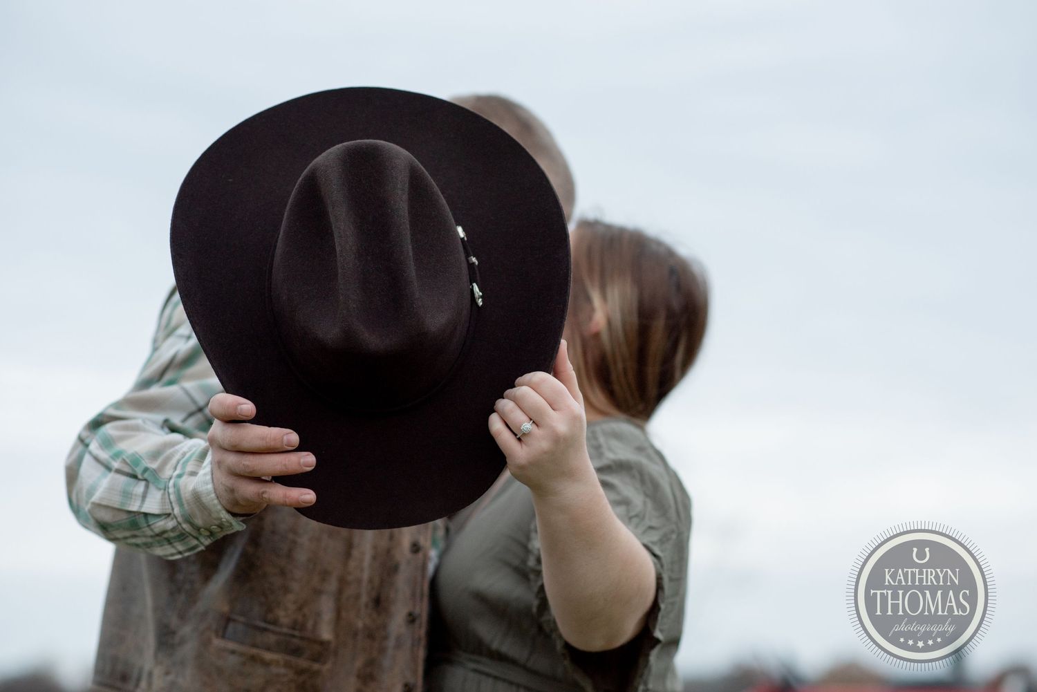 A creative photo showing a couple kissing behind a brown cowboy hat against an overcast sky.