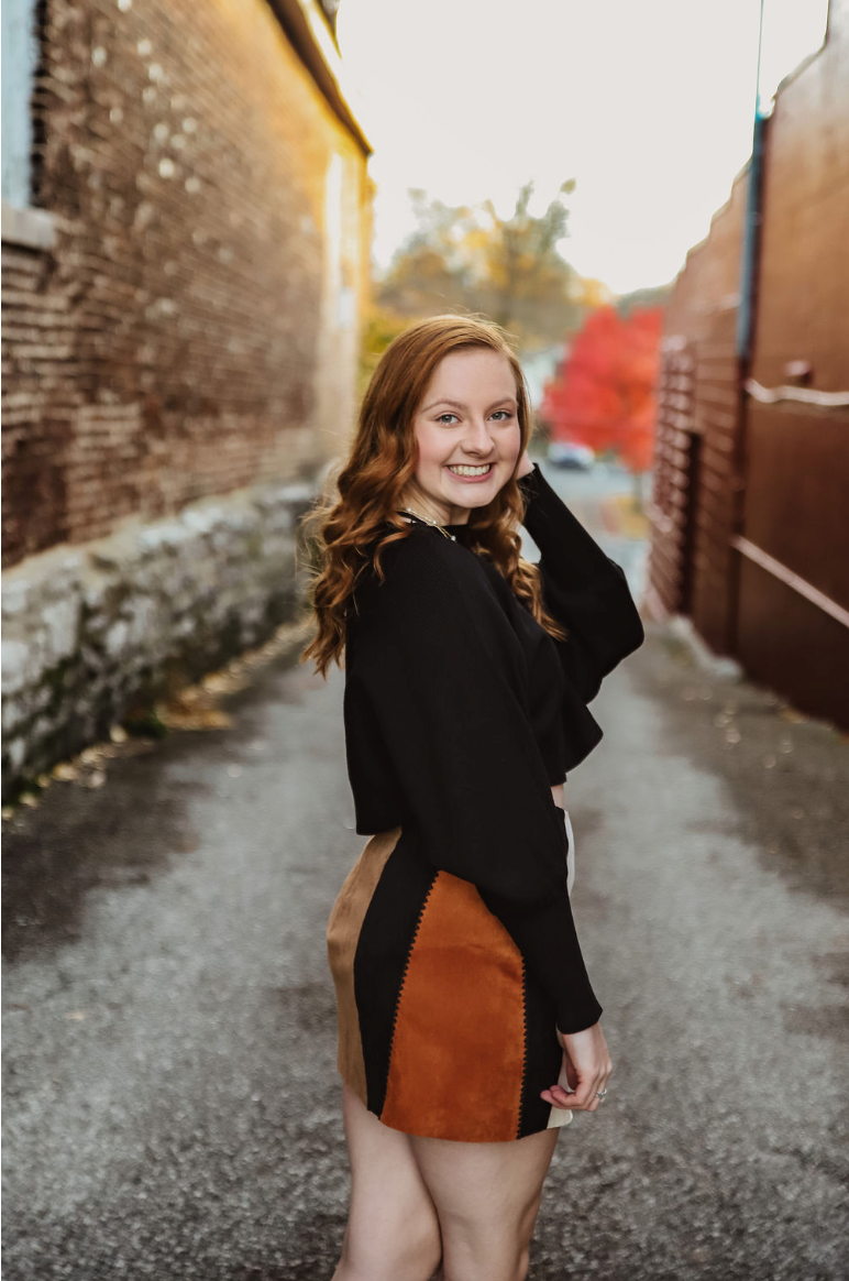 Portrait of a woman in black sweater and brown skirt against brick wall at sunset.