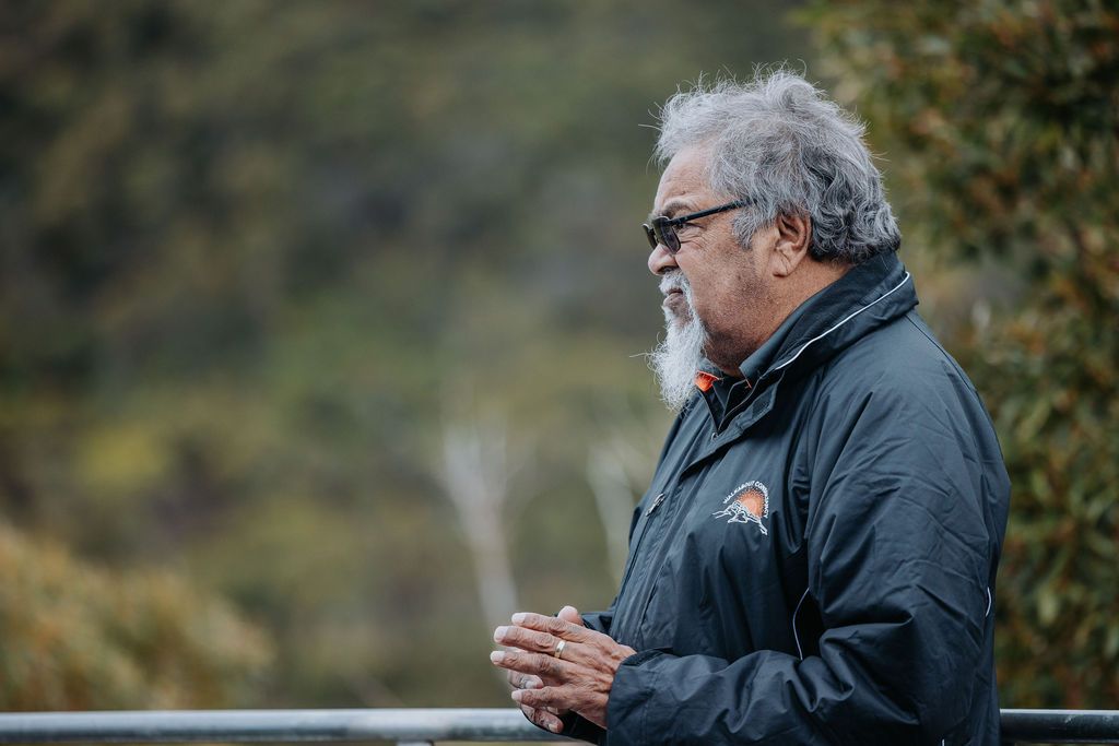 A person in a black jacket gestures while speaking to an attentive outdoor audience.