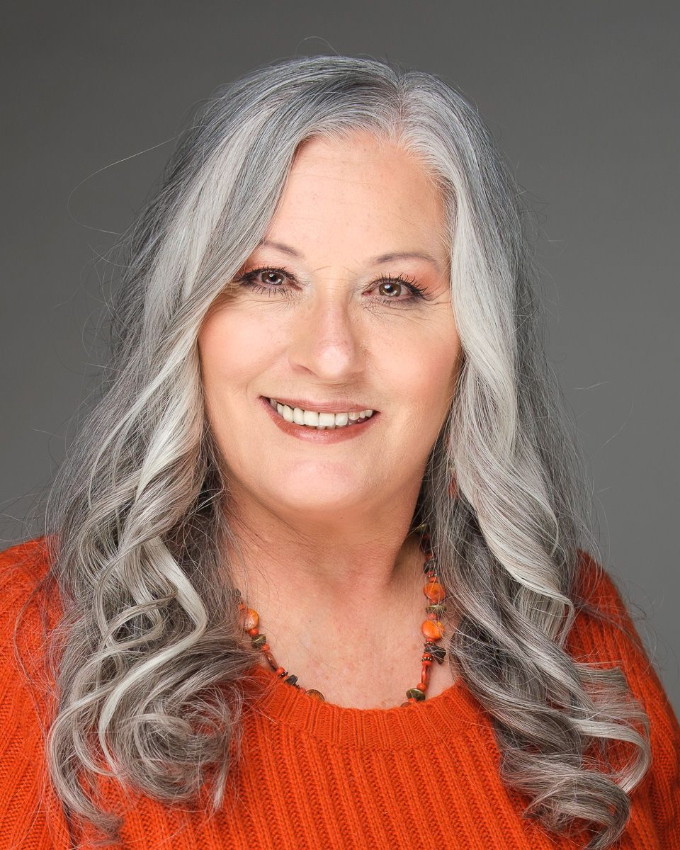 Professional portrait of someone with long wavy silver hair wearing an orange sweater and beaded necklace against gray background.