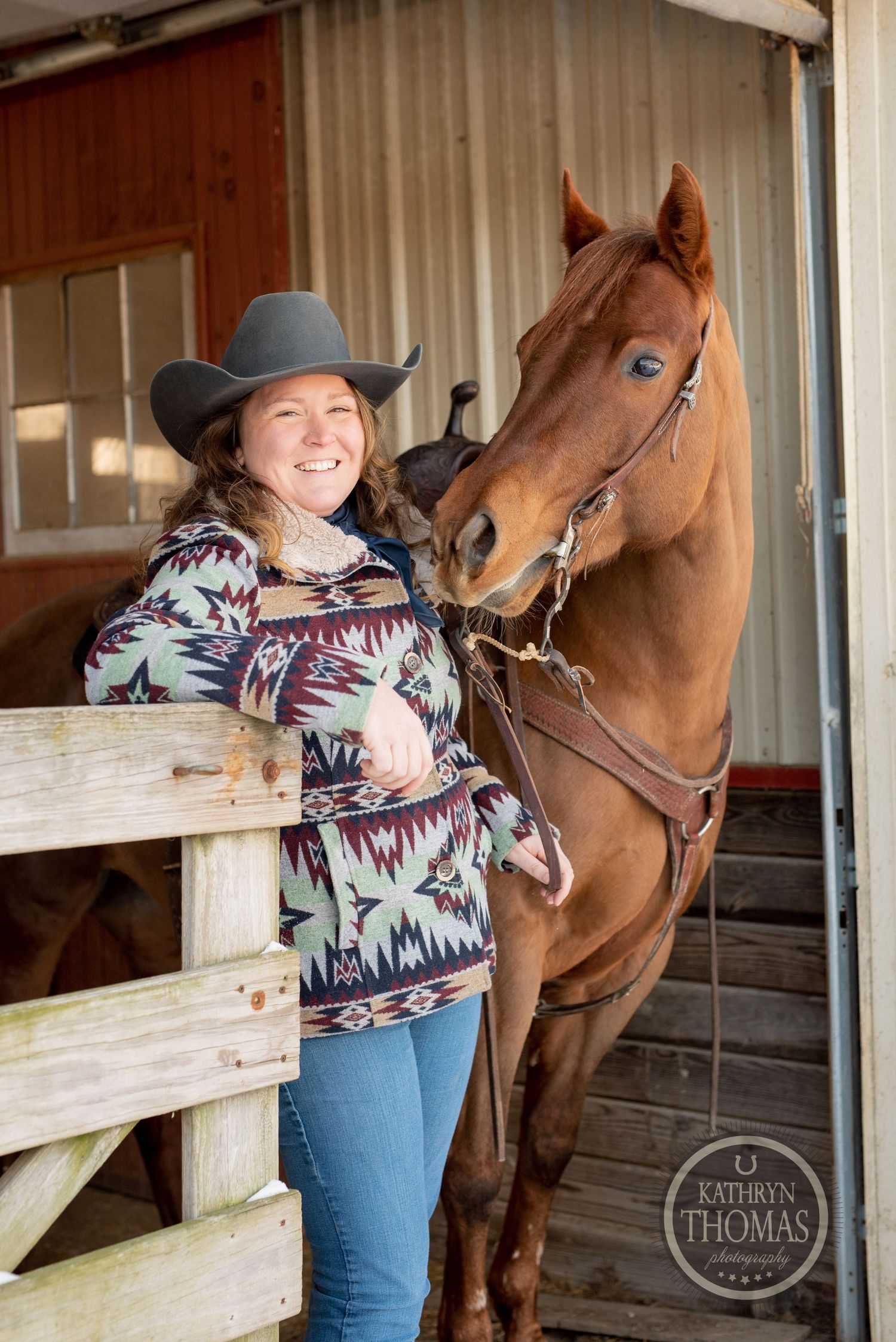 Brown horse and handler in cowboy hat and southwestern print sweater at wooden stable entrance.